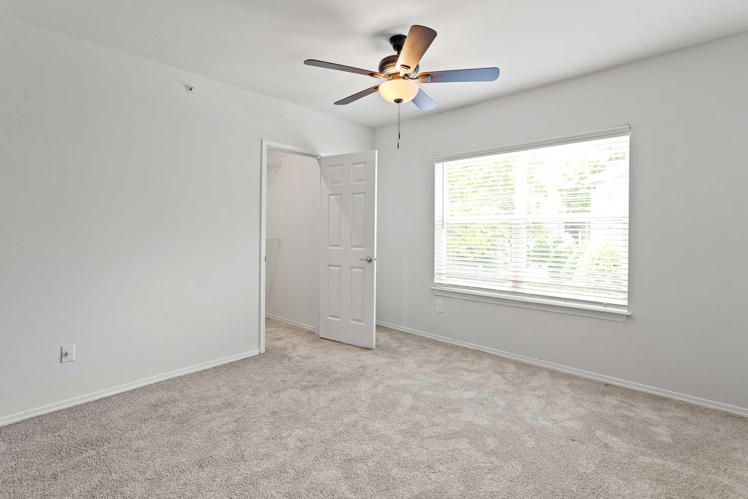 A spacious, empty room with light-colored walls and carpet. There is a ceiling fan with four blades and a light fixture. A window provides natural light, featuring white vertical blinds. An open door leads to a closet area, enhancing the sense of openness in the room.