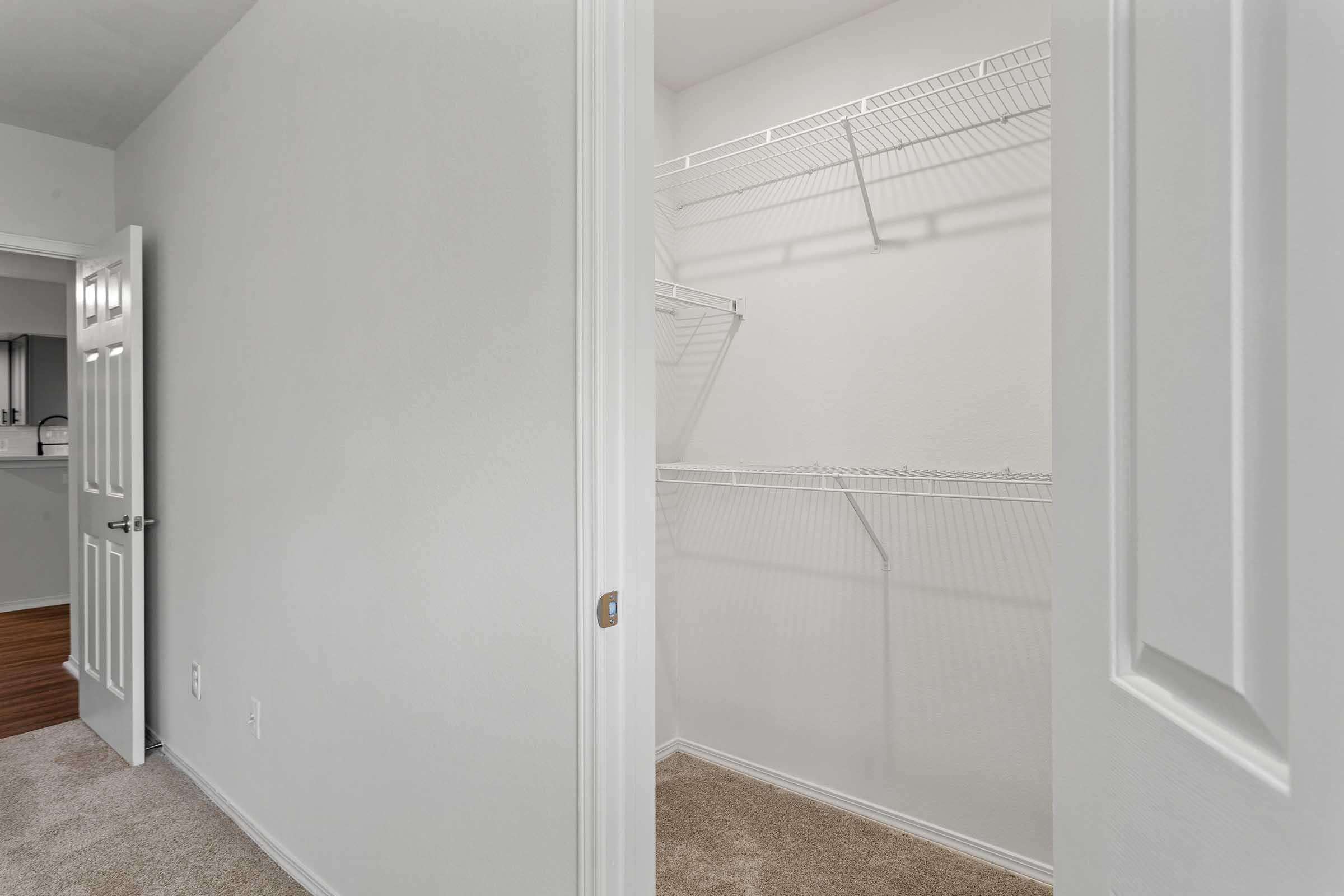 A view of a partially open closet with empty wire shelving, featuring light-colored walls and carpeted flooring. The doorway to the closet is in focus, with a neutral color scheme creating a spacious feel. In the background, a glimpse of a kitchen area can be seen.
