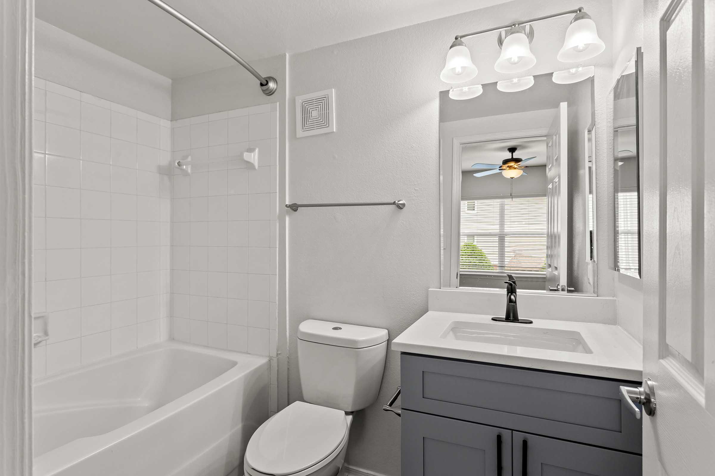 A clean and modern bathroom featuring a shower and bathtub combo, a white toilet, and a stylish gray vanity with a sink. The space is well-lit by overhead lights and has a mirror reflecting a window with blinds, enhancing the bright and airy feel of the room.