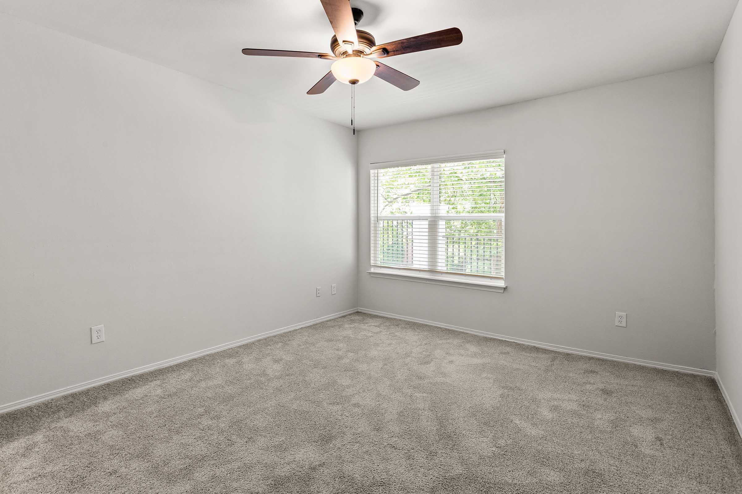 A bright, empty bedroom featuring light gray walls and a ceiling fan. The room has soft beige carpet flooring and a large window with natural light coming through, providing a view of greenery outside. The space is uncluttered, creating a fresh and airy atmosphere.