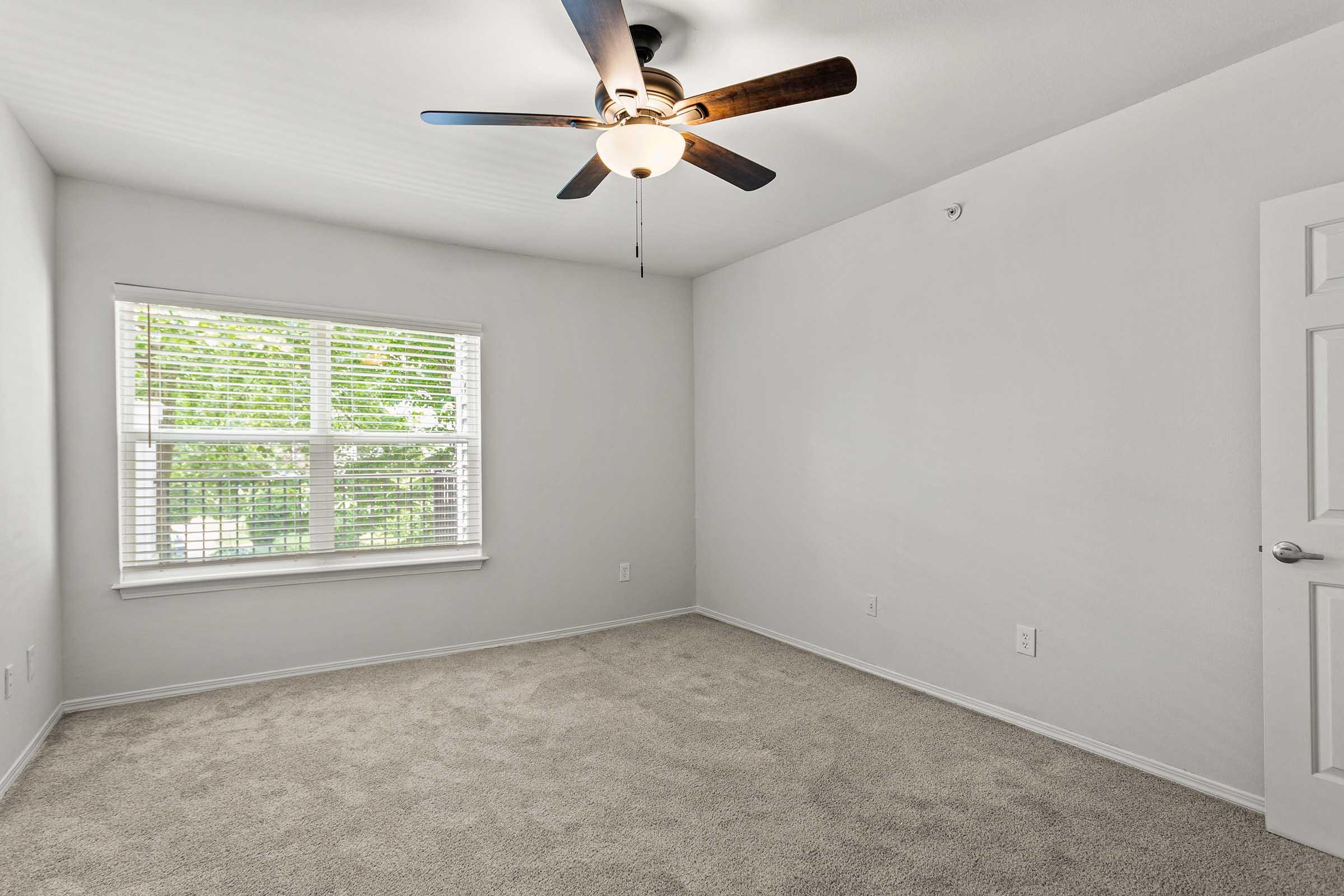 A bright, empty room featuring light-colored walls and plush carpet. A ceiling fan hangs from the ceiling, and a window with white blinds allows natural light to enter, illuminating the space. The room is unadorned and ready for personalization.