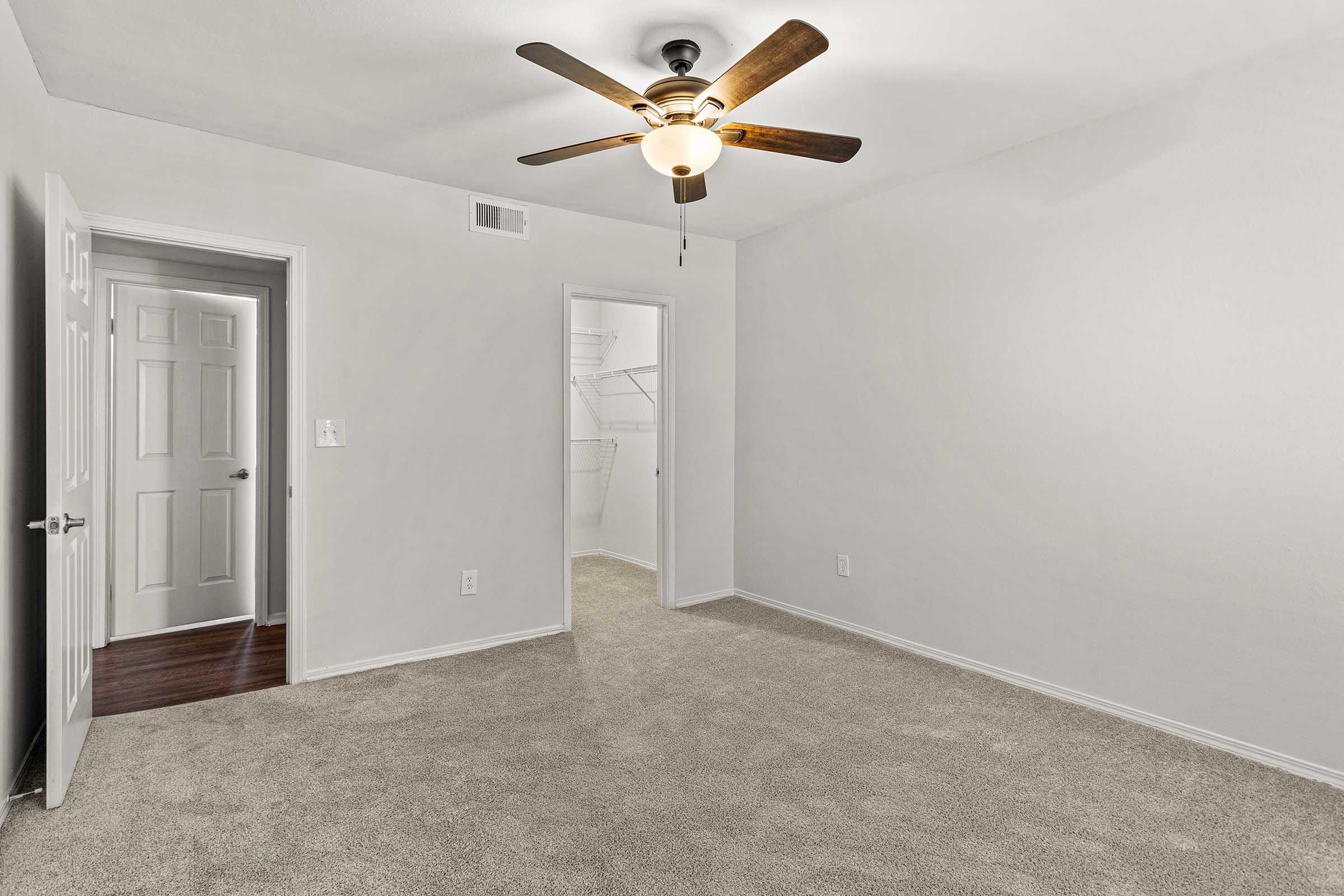 Empty room with light gray walls and tan carpet. A ceiling fan with wooden blades hangs from the ceiling. Two doors are visible, one leading to a closet and the other to a hallway. Natural light is minimal, creating a simple and clean aesthetic. No furniture or decor present.
