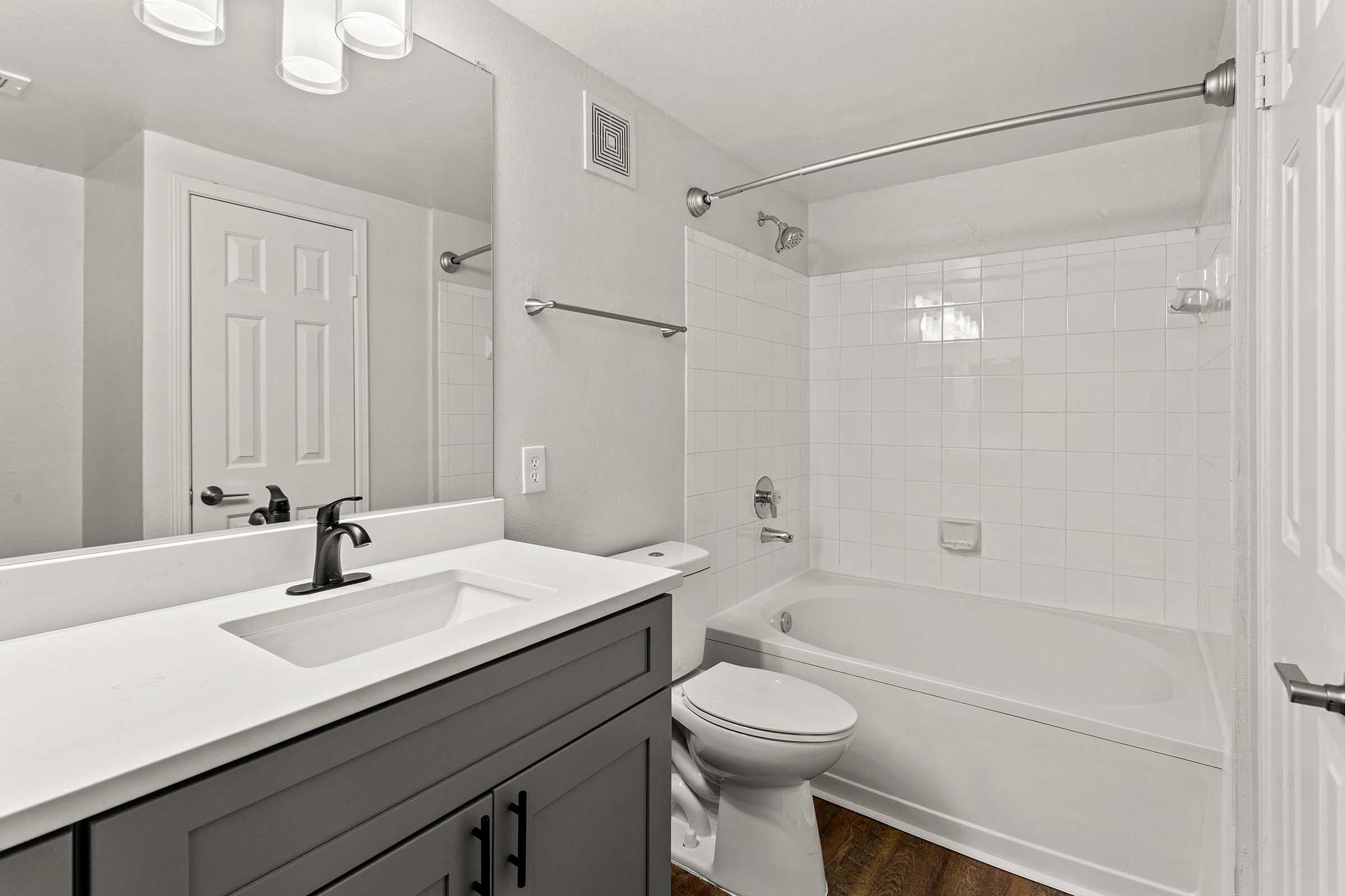 A modern bathroom featuring a white tiled bathtub and shower combination, a sleek white countertop with a black faucet, a toilet, and a mirrored vanity. The walls are painted in neutral tones, and there is a door leading out of the space, adding to the clean and contemporary design.