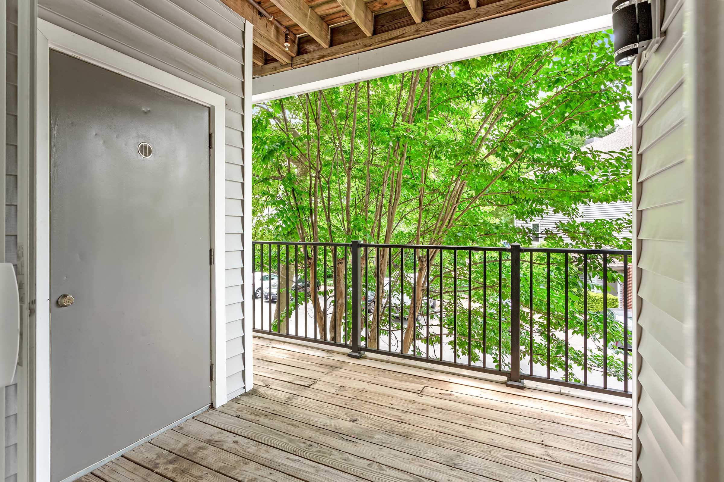 A wooden balcony with a railing overlooks a lush, green tree outside. There is a plain gray door on one side, and the surrounding area is well-lit by natural sunlight, creating a serene and inviting atmosphere.