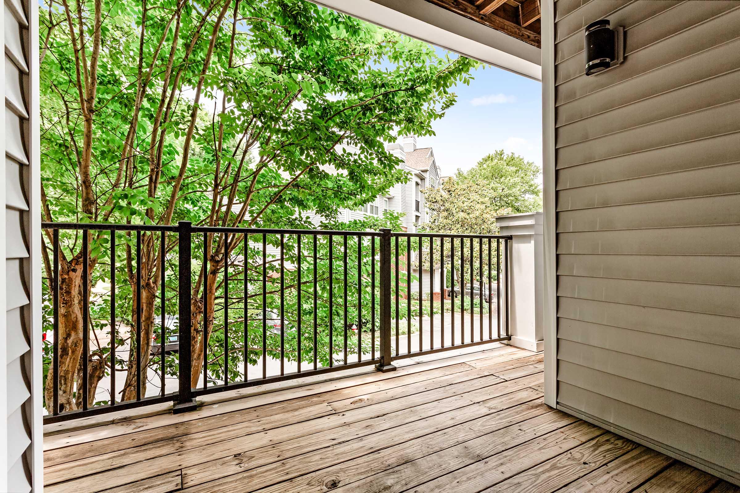 A view of a wooden balcony with a black railing, overlooking a green tree and residential buildings. The sky is clear with a few clouds, creating a bright and inviting atmosphere. The balcony is partially shaded by the overhanging roof above.