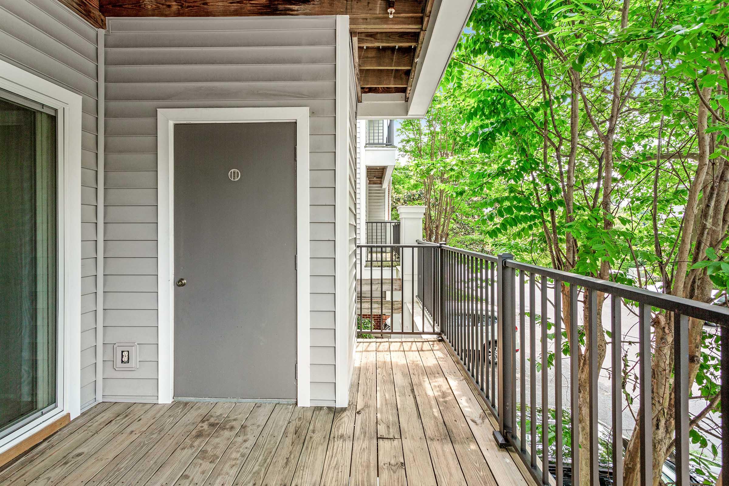 A balcony with wooden flooring, featuring a gray door labeled "11." Lush green trees are visible, adding a natural touch to the space. The house exterior has gray siding, and there is a railing along the balcony's edge. Sunlight filters through the leaves, creating a bright and inviting atmosphere.