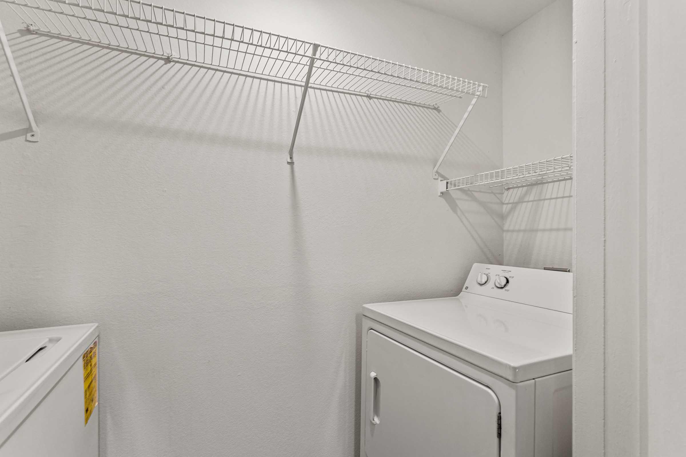 A clean, small laundry room featuring a white washer and a dryer side by side. Above, a wire shelf is mounted on a plain white wall, providing storage space. The room is well-lit and organized, emphasizing a tidy and functional laundry area.