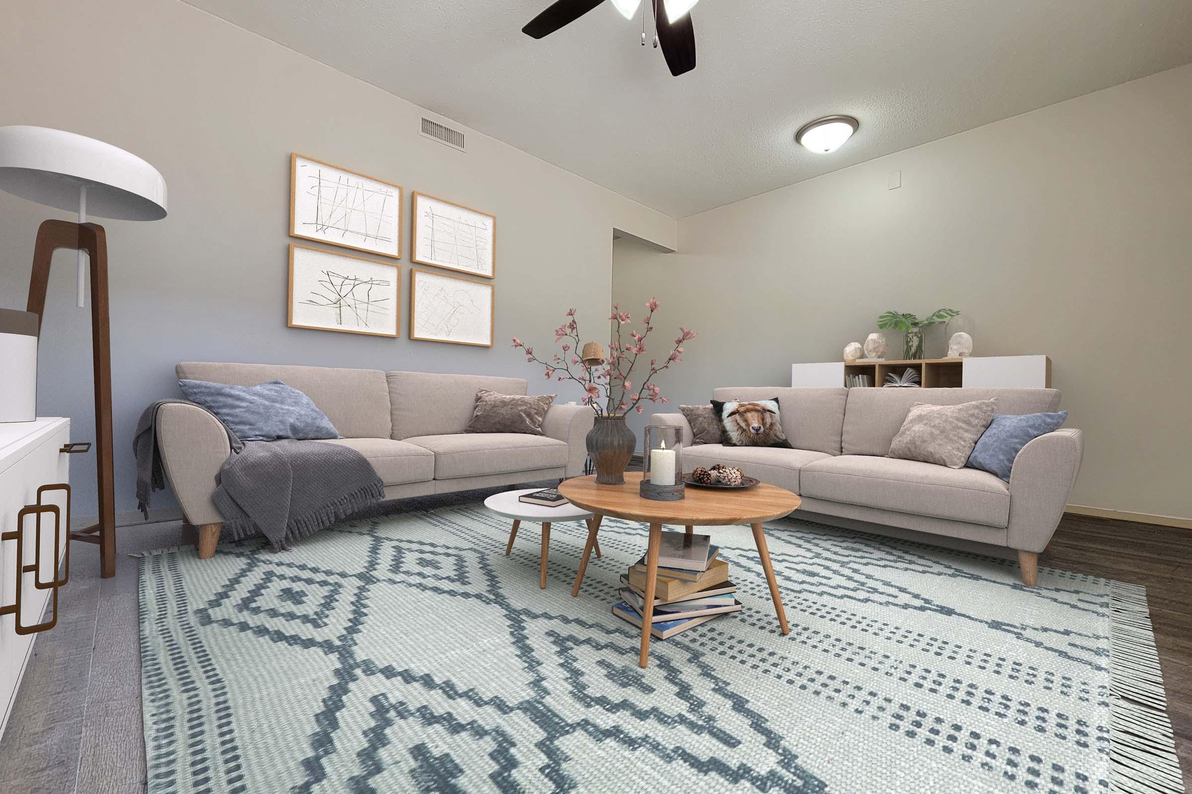 A cozy living room featuring a light gray sofa with pillows, a circular wooden coffee table on a patterned blue area rug, and modern decor including a vase with flowers and framed artwork. A standing lamp and a white cabinet are also visible, along with neutral-toned walls and a ceiling fan.