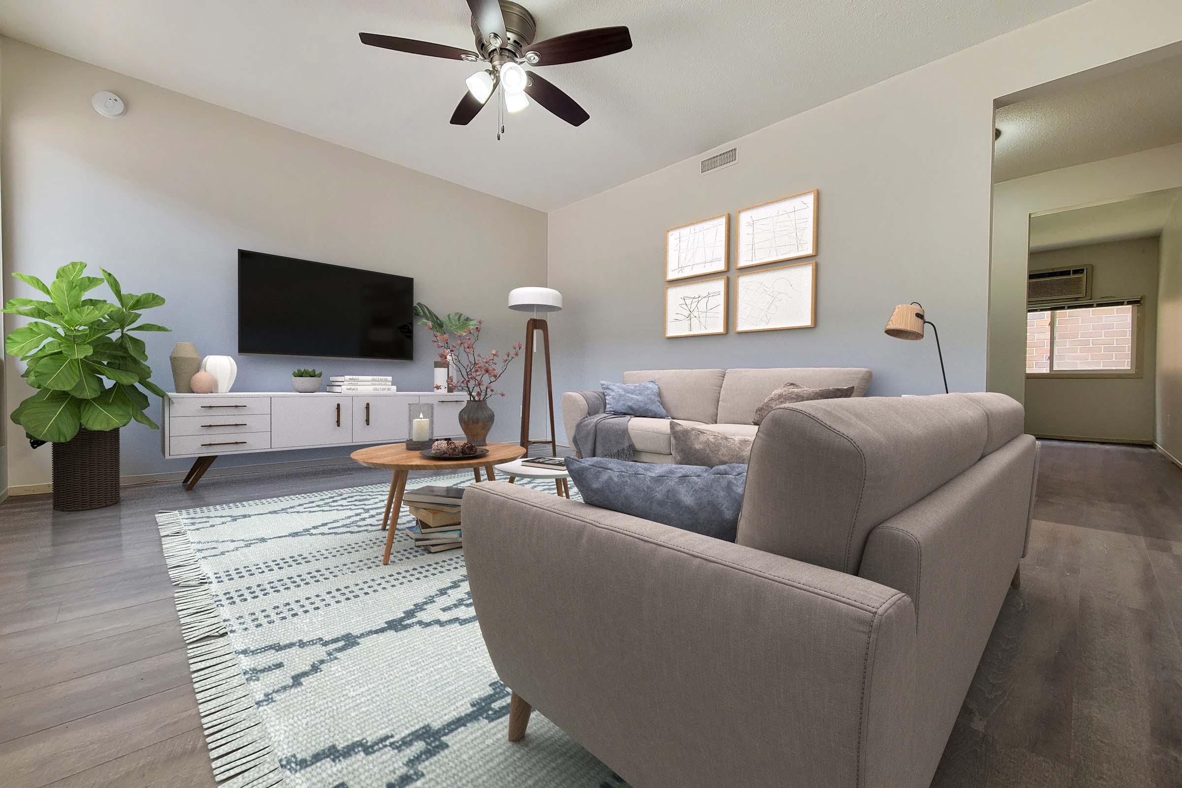 A modern living room featuring a light gray sofa, decorative coffee table, and a large flat-screen TV mounted on the wall. A potted plant adds a touch of greenery, while three framed artworks hang on the wall. The space has a neutral color palette with a patterned area rug on the wooden floor, creating a cozy atmosphere.