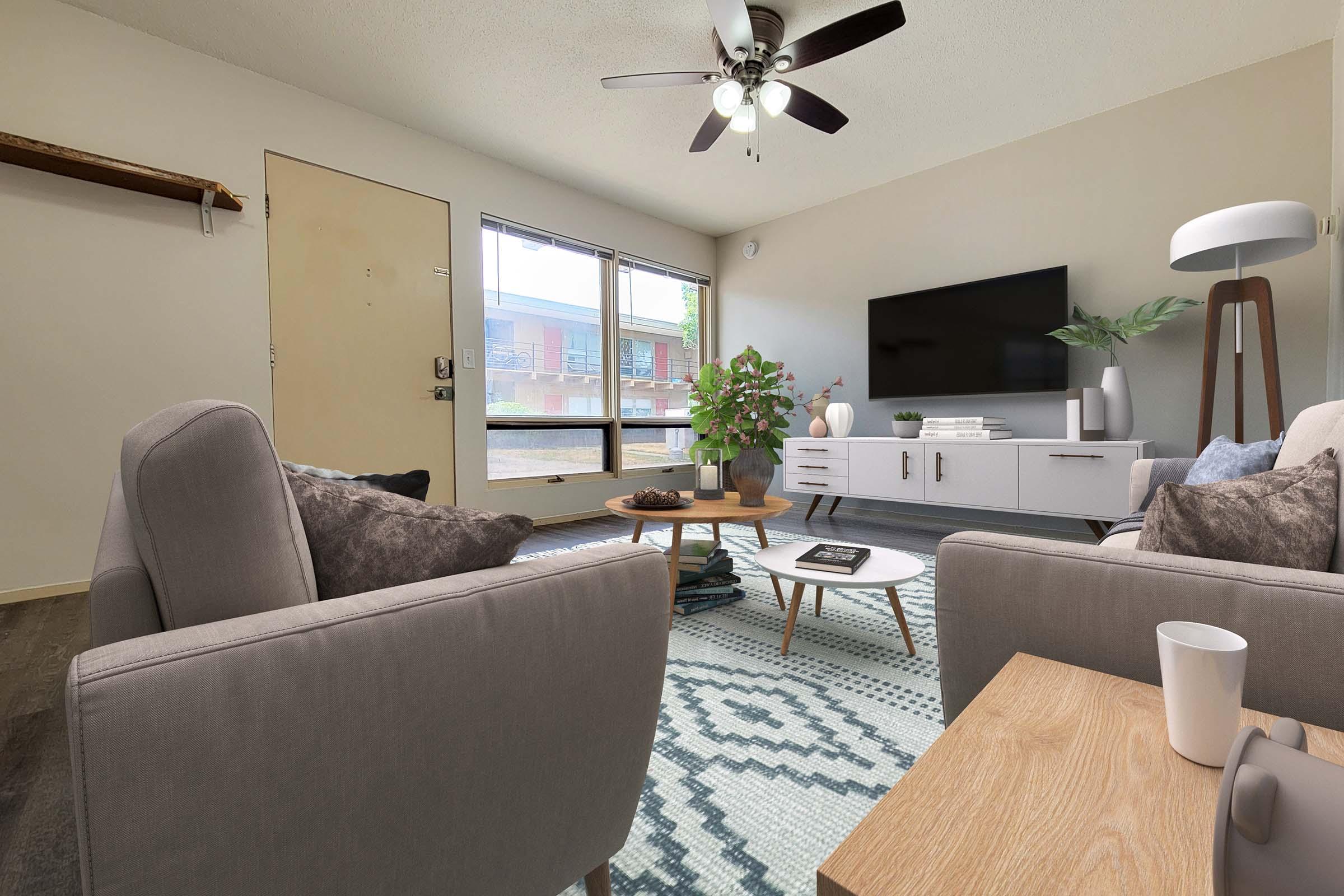 A cozy living room featuring two gray sofas with decorative pillows, a round wooden coffee table, and a stylish TV stand with a flat-screen TV. A ceiling fan hangs above, and a large window provides natural light. A potted plant and a small side table add finishing touches to the inviting space.