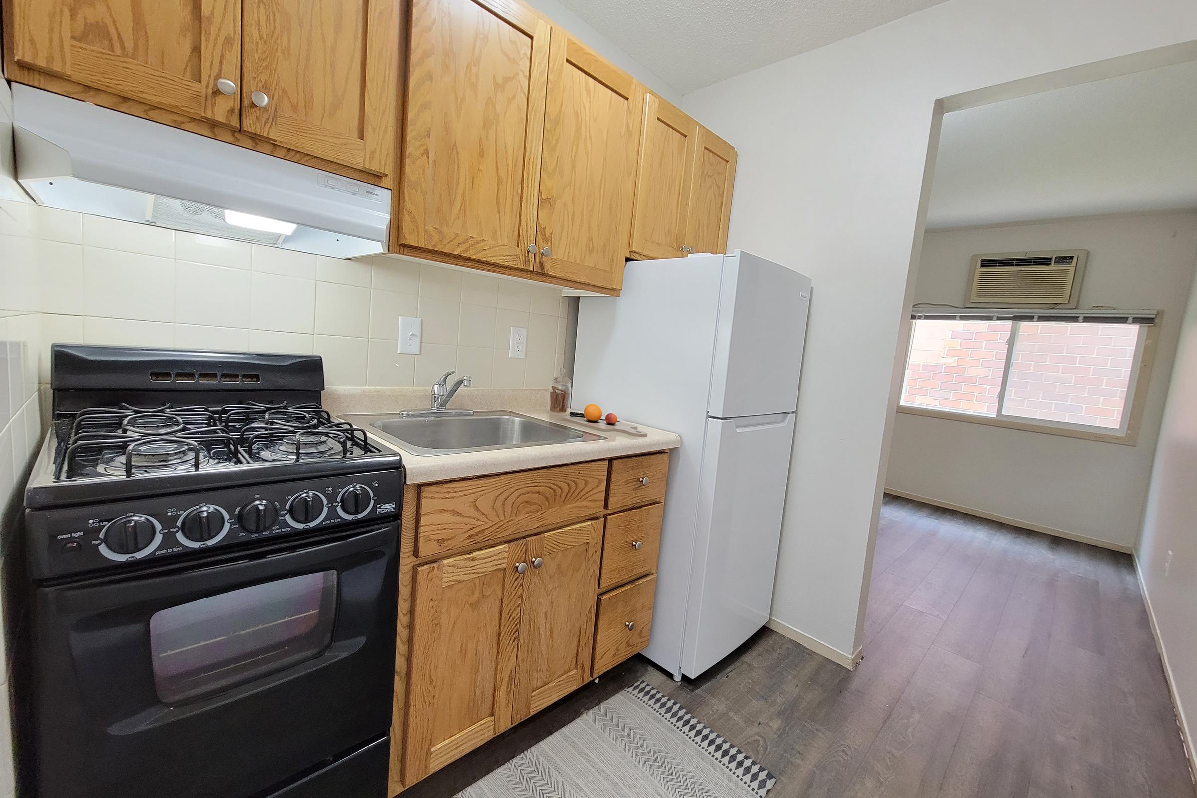 Small kitchen with wooden cabinets, a gas stove, and a sink. A white refrigerator is visible next to the sink. There is a window in the background leading to a bright room with wooden flooring. An orange is placed next to the sink. The space is well-lit and organized, featuring a simple layout.