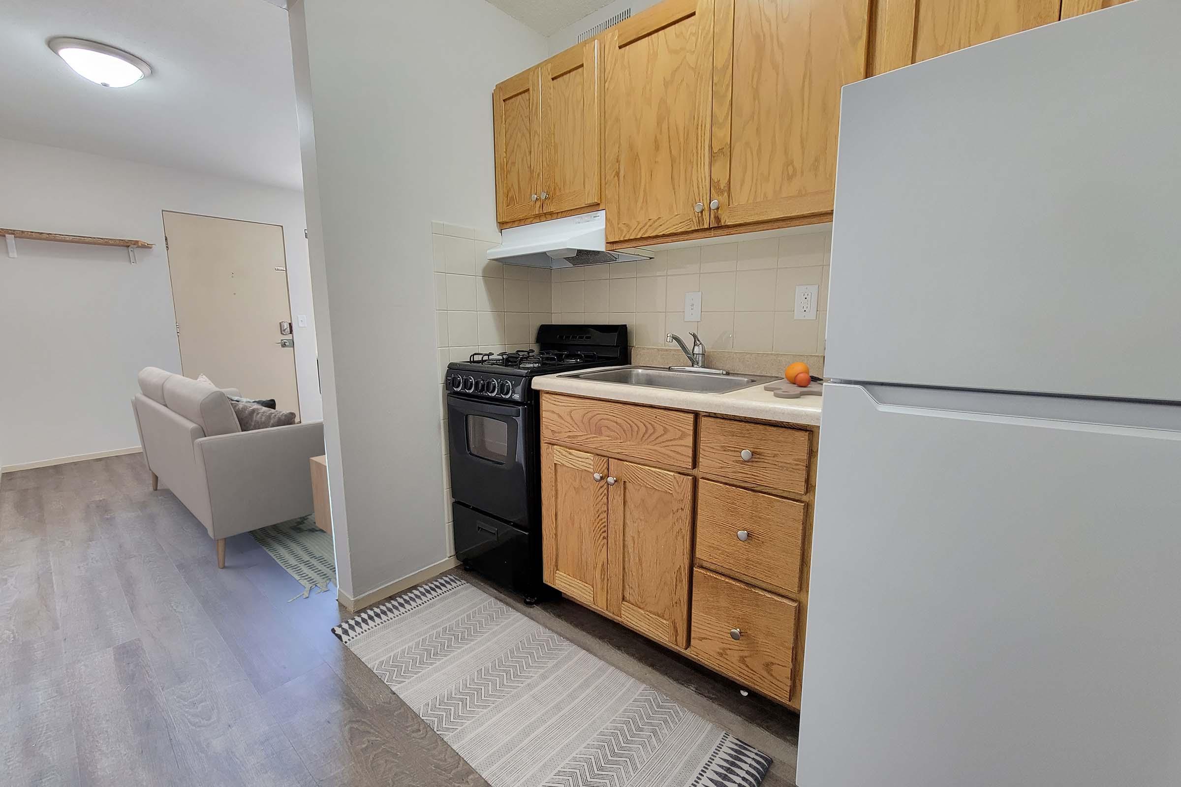 A small kitchen area featuring wooden cabinets, a stove, a sink, and a refrigerator. The kitchen is part of an open layout that connects to a living space with a light gray sofa in the background. The floor has a light wood finish, and a patterned rug runs in front of the kitchen appliances.