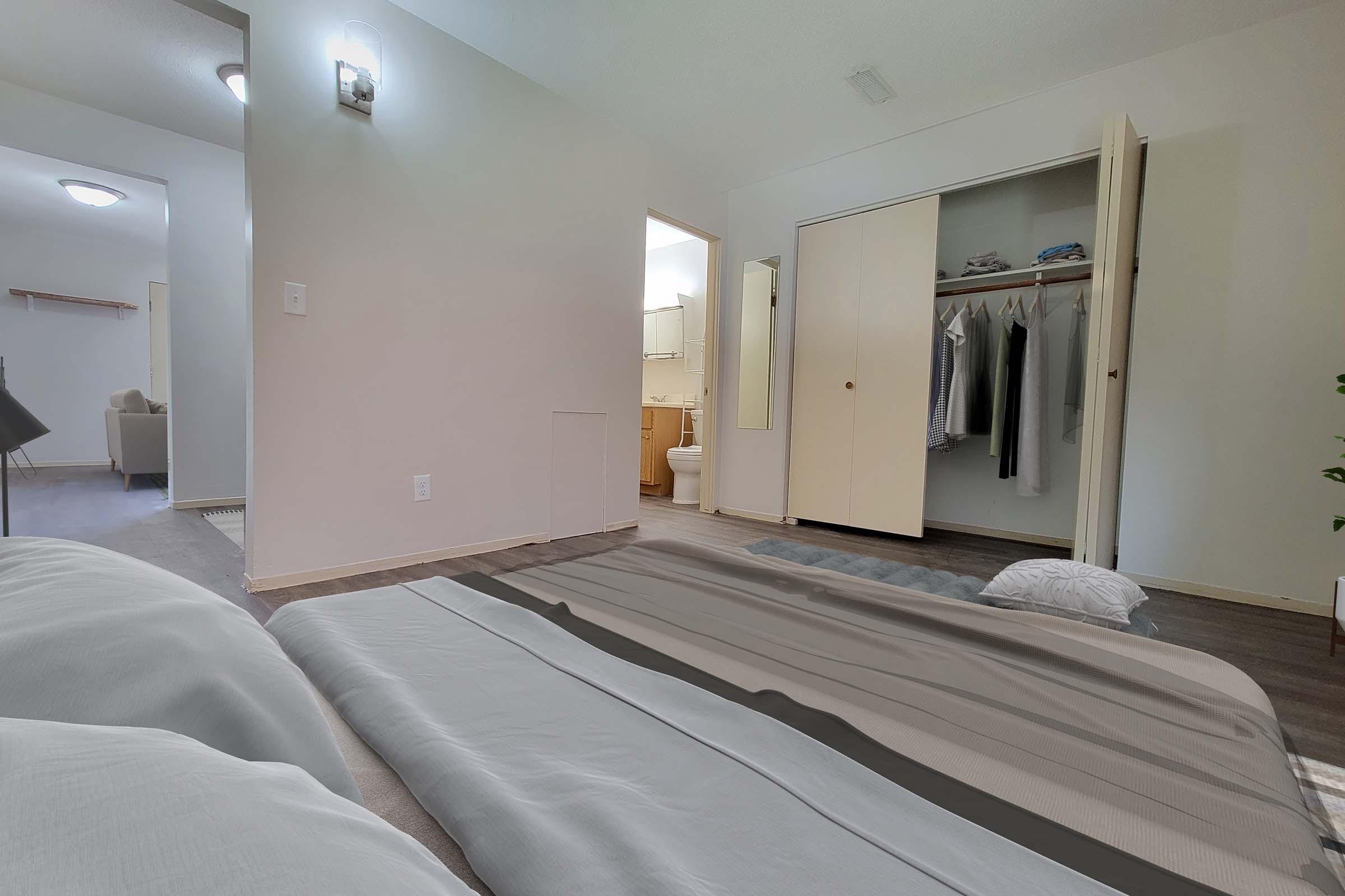 A minimalist bedroom setup with bedding laid on the floor. The room features a closet with sliding doors, an en-suite bathroom visible in the background, and soft lighting. Neutral color tones and simple decor create a calm atmosphere.
