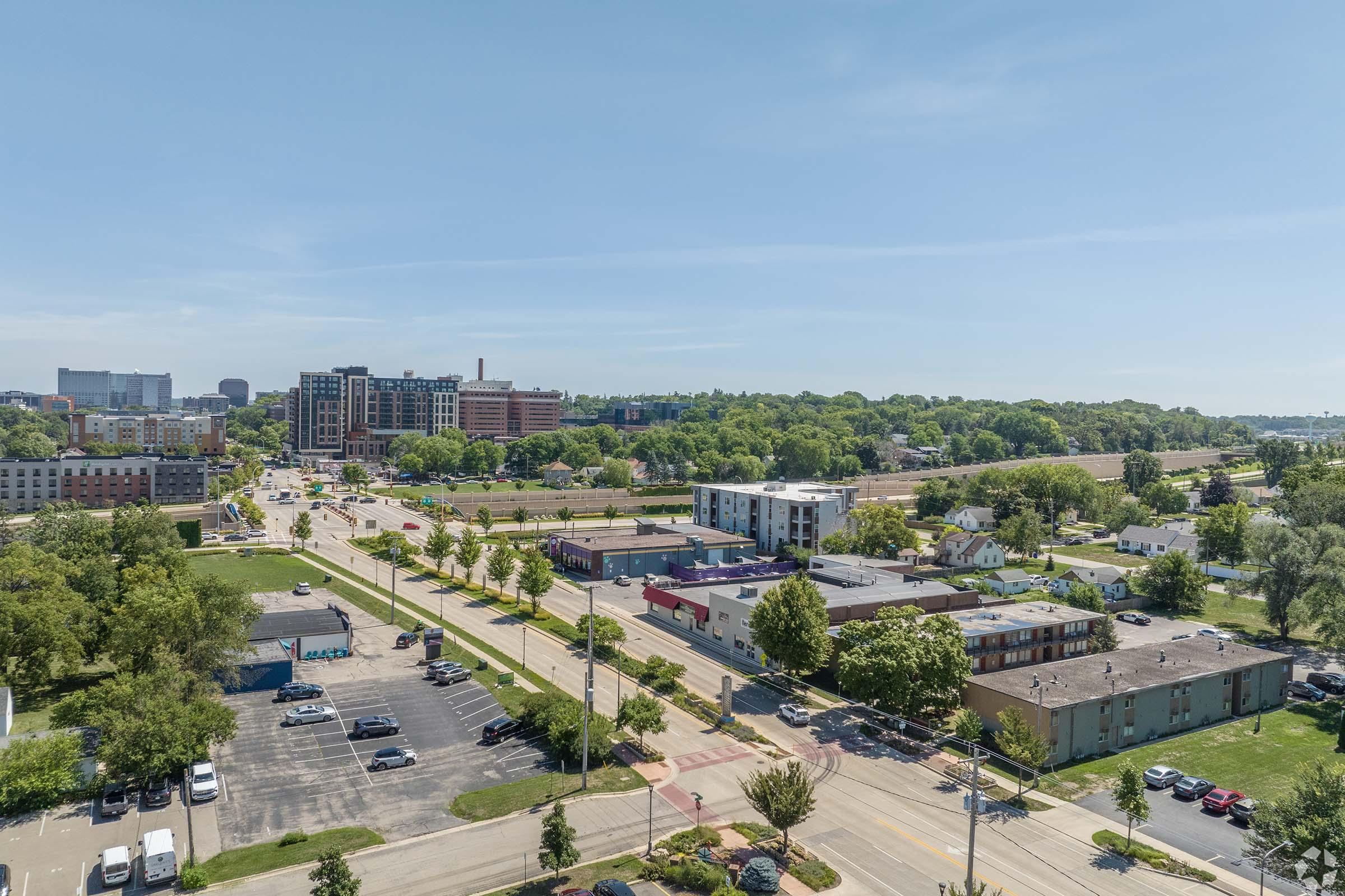 Aerial view of a cityscape featuring a mix of residential and commercial buildings, green trees, and an open area. The scene includes a road lined with trees and several parked cars in the foreground, with taller buildings and a blue sky in the background.