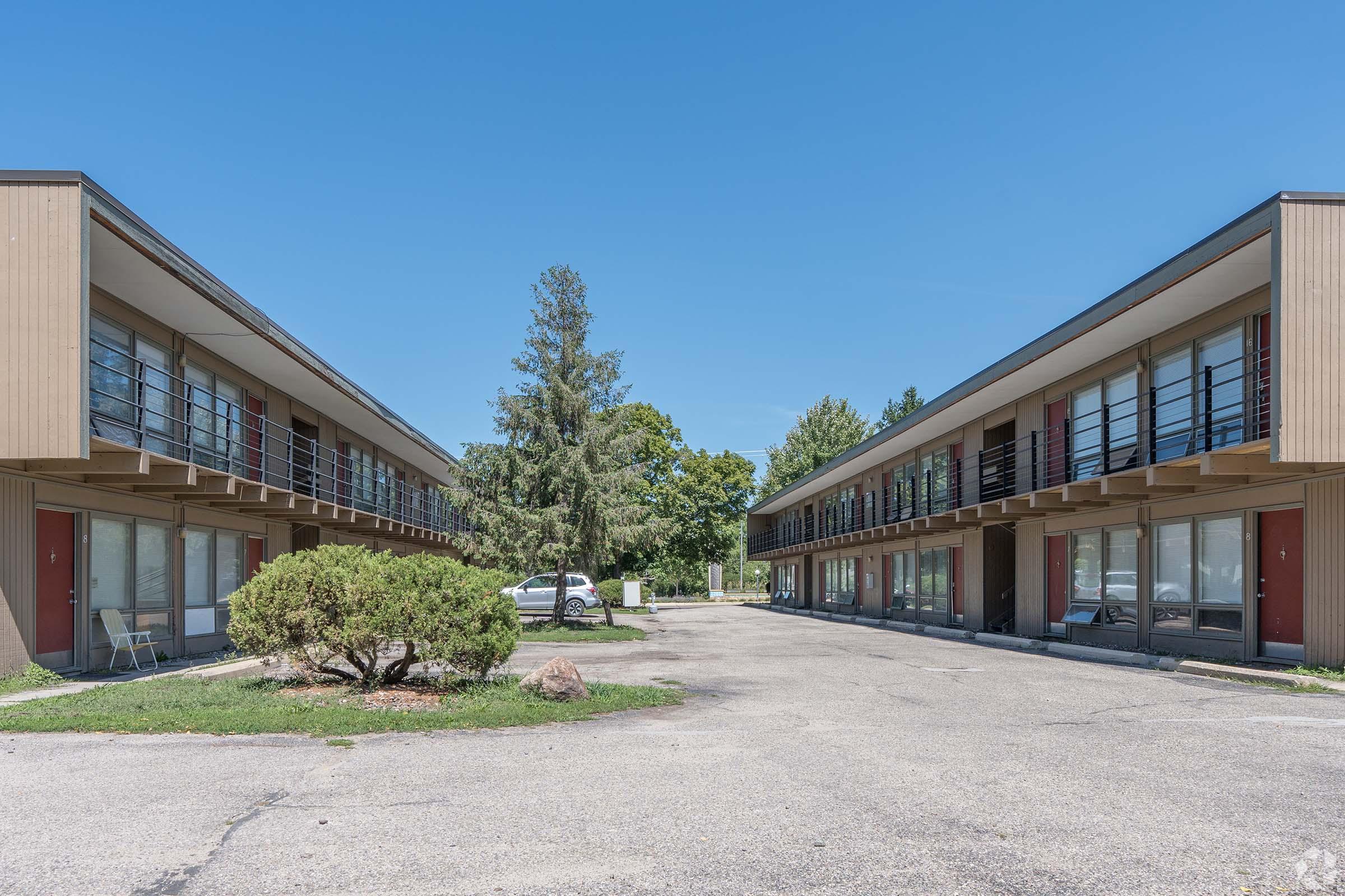 A view of a two-story motel with balconies on both sides, lined up along a paved driveway. The area features greenery, including a small bush and trees in the background, under a clear blue sky. The parking lot is empty, creating a tranquil ambiance around the motel.