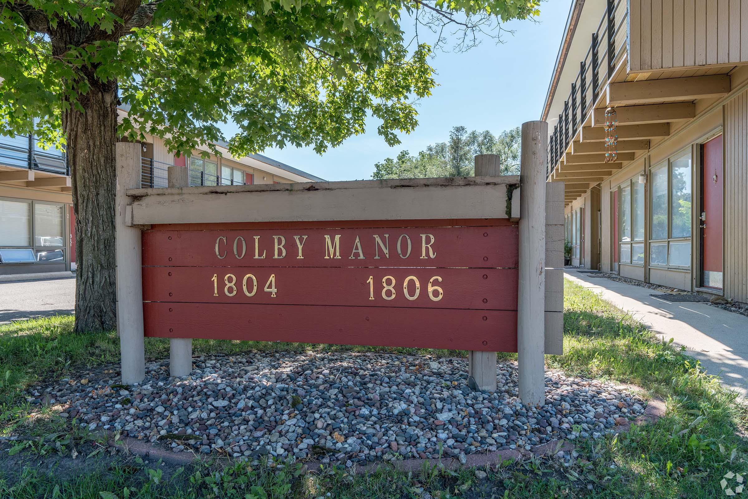 Sign for Colby Manor displaying the numbers 1804 and 1806, set amidst grass and small stones, with a tree nearby. The building features a simple architectural style with multiple entrances visible.