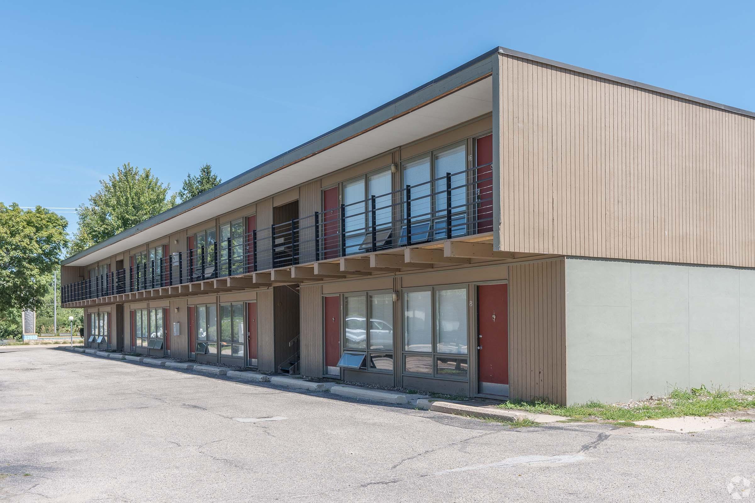 A two-story motel with a modern design featuring a flat roof, multiple balconies, and red doors. The building is situated in an empty parking lot surrounded by trees and clear blue sky. Some windows are visibly open, suggesting occupancy. The overall ambiance is quiet and somewhat deserted.