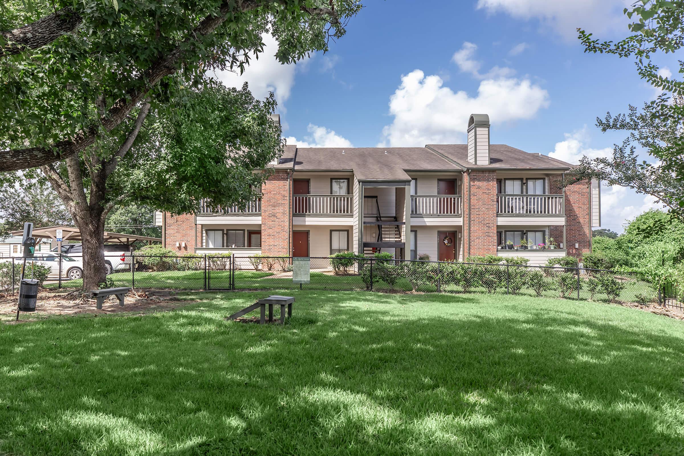 A well-maintained residential building with brick exterior and balconies, surrounded by green grass and trees. The scene features a clear blue sky with fluffy clouds, showcasing a peaceful and inviting atmosphere. Parking spaces are visible nearby, indicating an accessible living environment.