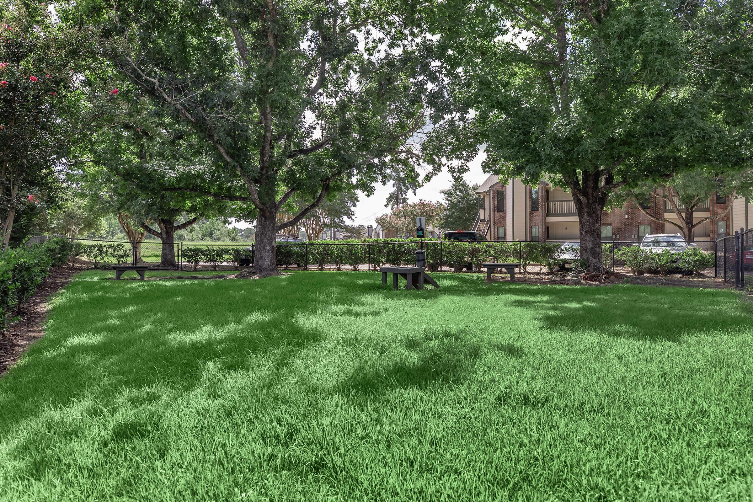 A shaded green park area with two large trees and a bench, surrounded by neatly trimmed grass. In the background, there are buildings and a parking lot visible, providing a serene outdoor space.