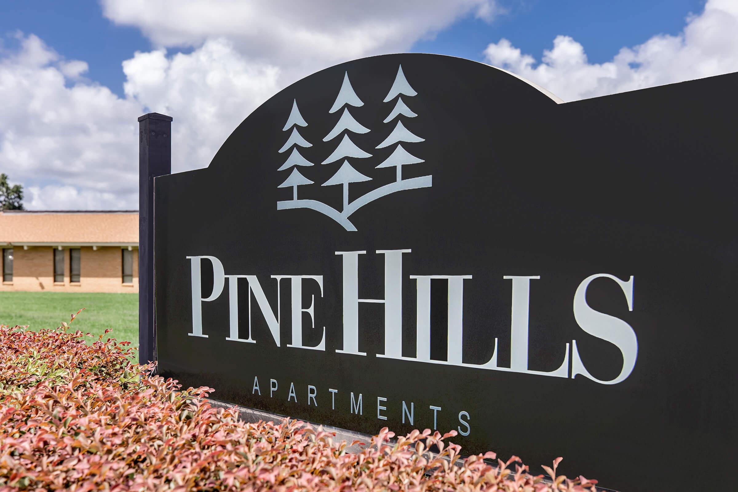 Sign for Pine Hills Apartments featuring stylized trees and a clear blue sky in the background. The sign is prominently displayed with the text "PINE HILLS" and "APARTMENTS" beneath, set against a decorative border. Green grass and colorful shrubbery are visible at the base.