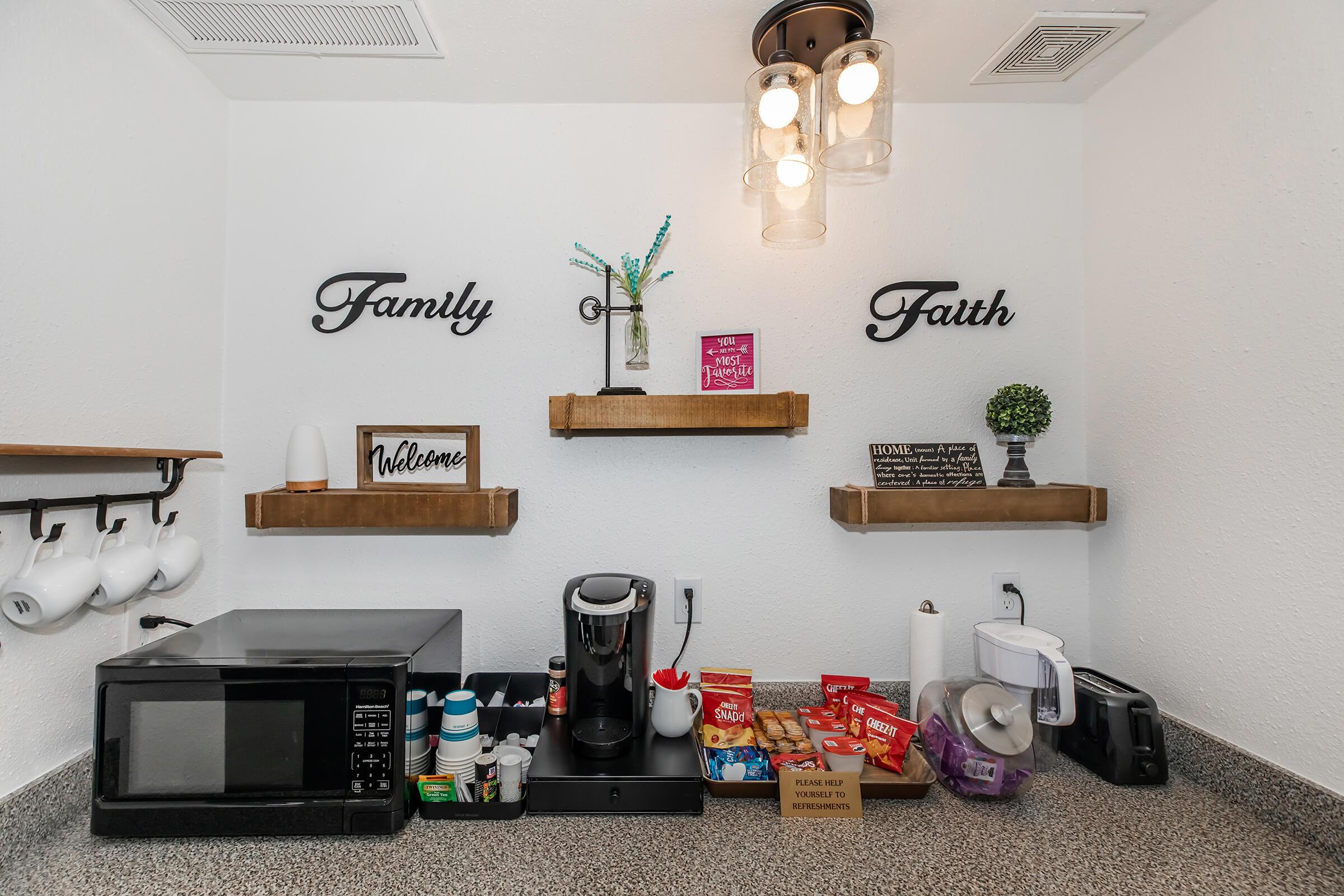 A cozy kitchen corner featuring shelves with decorative signs reading "Family" and "Faith." Below, there's a microwave, a coffee maker, and various snacks in boxes. The space has a welcoming feel, with a small plant and cups hanging on the wall. Warm lighting adds to the inviting atmosphere.