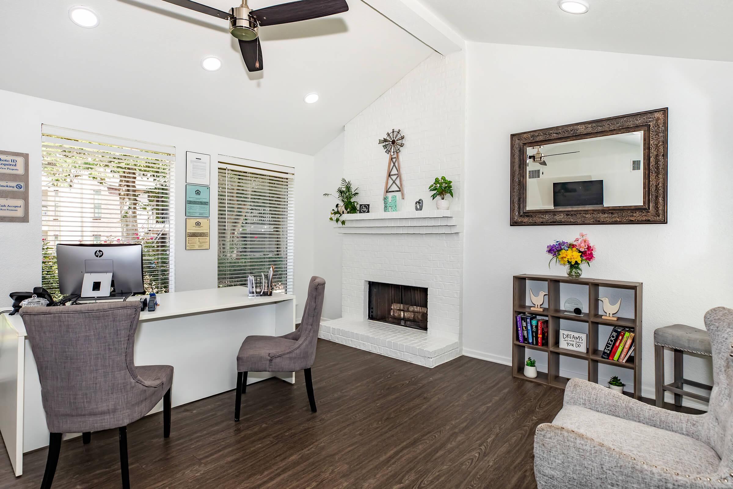 A bright and modern office space featuring a desk with a computer, two gray chairs, a fireplace with a decorative mantle, and a cozy seating area. Natural light filters through the windows, highlighting wall decorations and a mirror, creating an inviting and professional atmosphere.