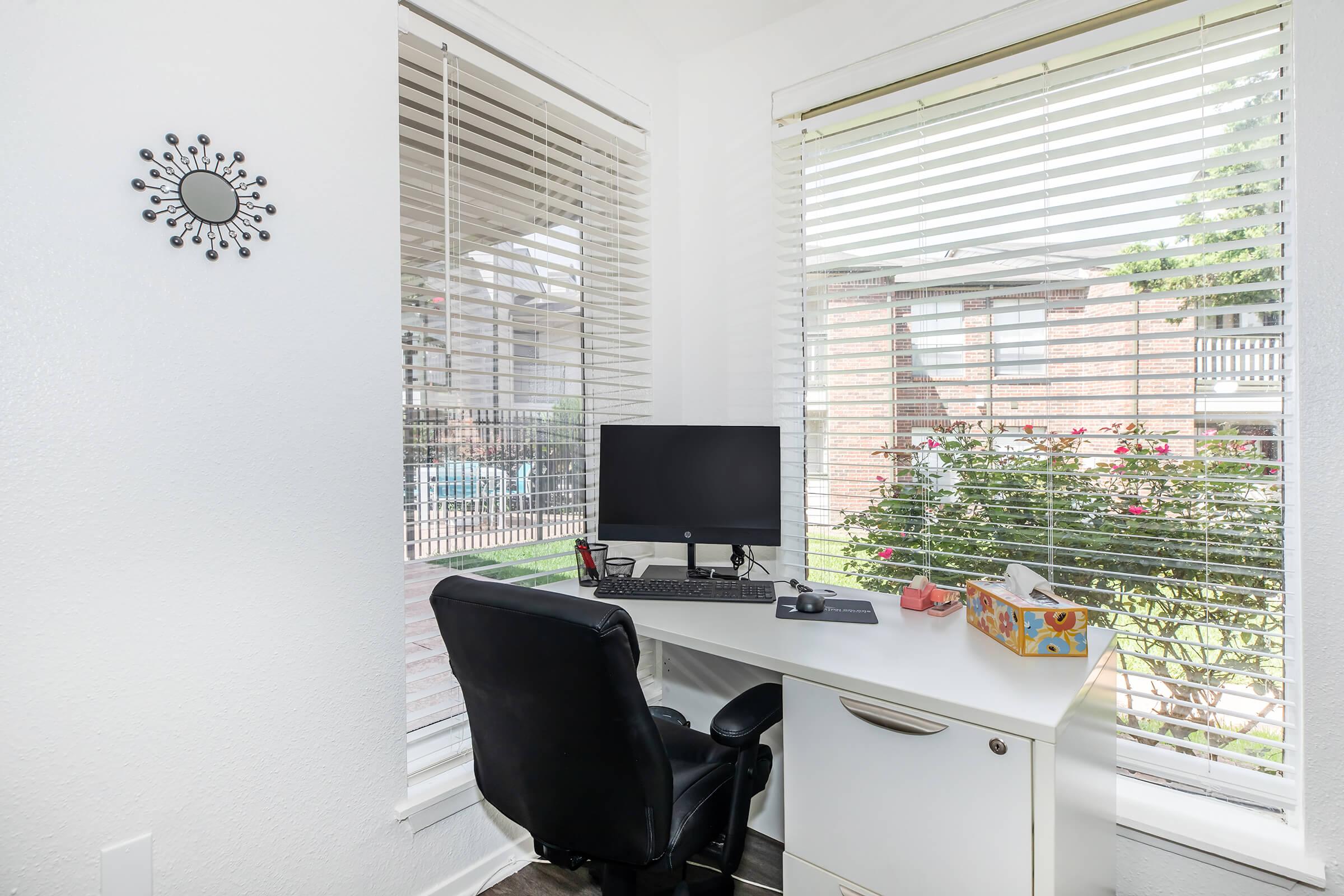 A bright home office with a white desk and a black ergonomic chair. A computer monitor is set up on the desk, facing two large windows with white blinds. Outside the windows, there are views of greenery and flowers. A small circular mirror is mounted on the wall.