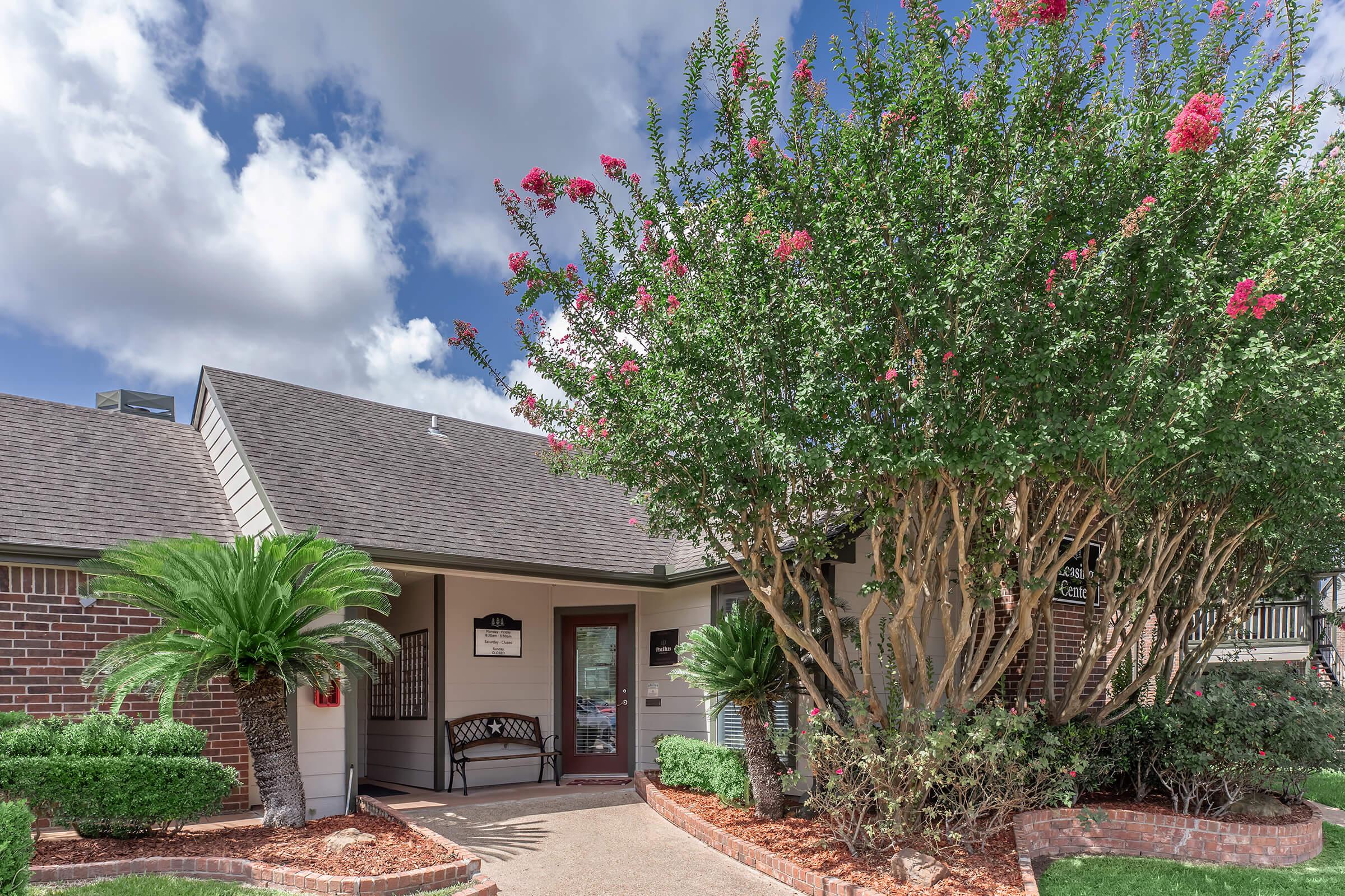 A charming entrance to a building featuring a covered porch, surrounded by lush greenery and flowering trees. The pathway leads to a door, with well-maintained landscaping including palms and colorful flowers, under a blue sky with fluffy white clouds.