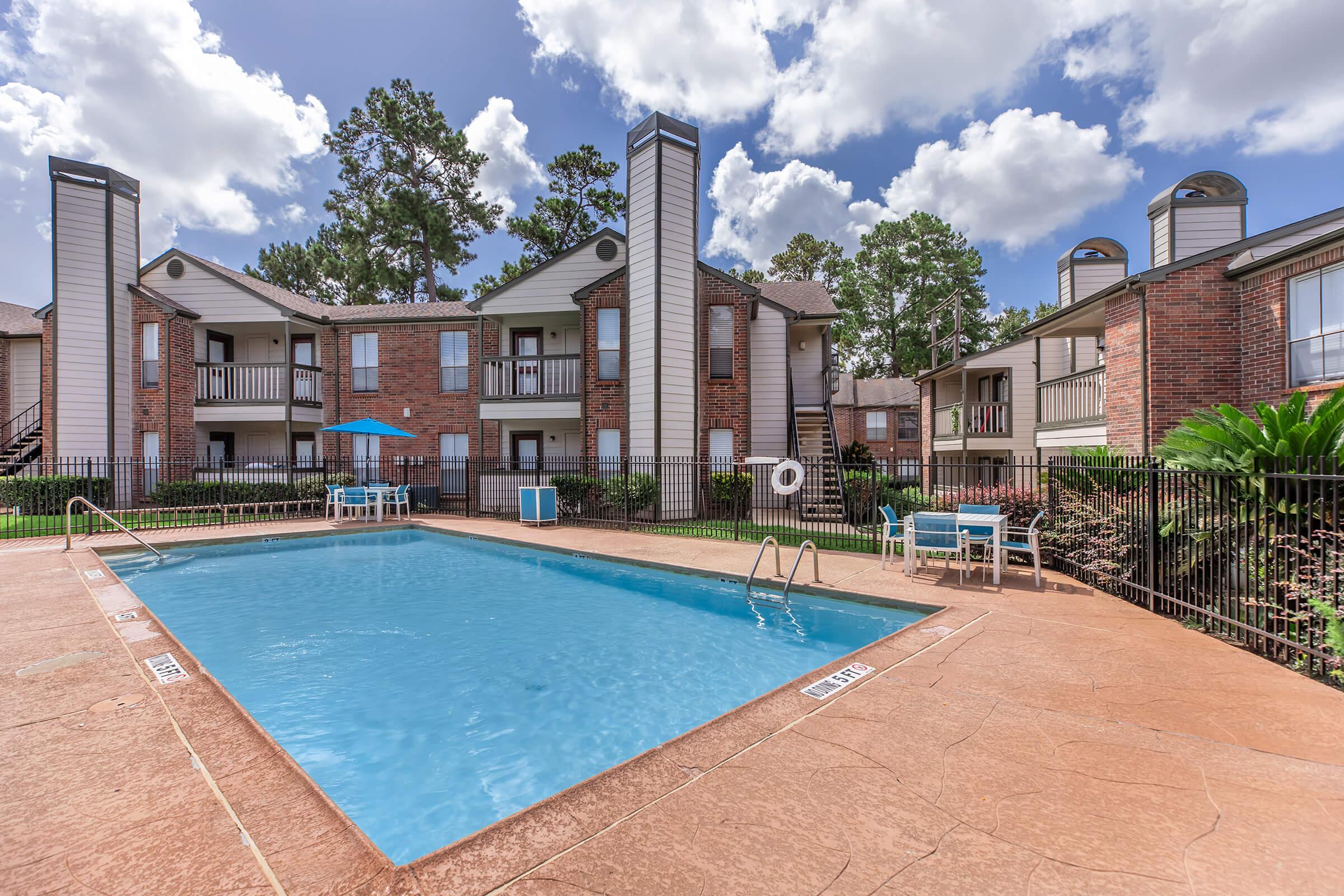 A clear blue swimming pool surrounded by a paved patio in an apartment complex. There are lounge chairs and an umbrella near the pool. The buildings are two stories with brick exteriors and balconies, set amidst trees and greenery under a partly cloudy sky.