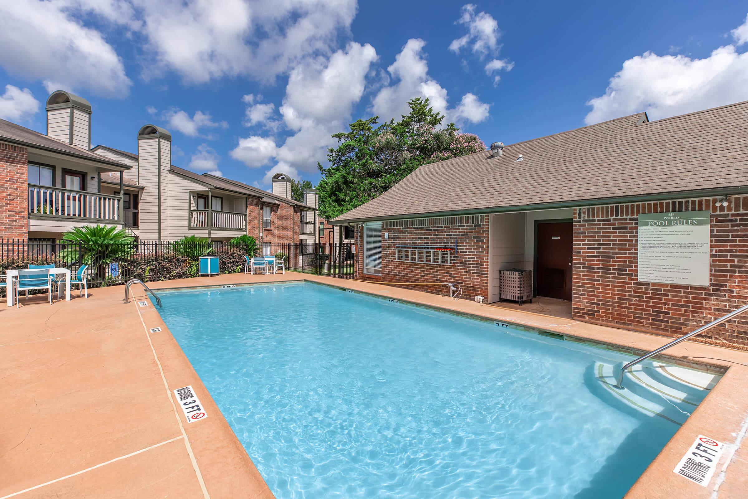 A sunny outdoor pool area surrounded by brick buildings with balconies, featuring lounge chairs and a small pool house. The pool is clear and inviting, with blue skies and fluffy white clouds above. Lush greenery adds a touch of nature to the setting.