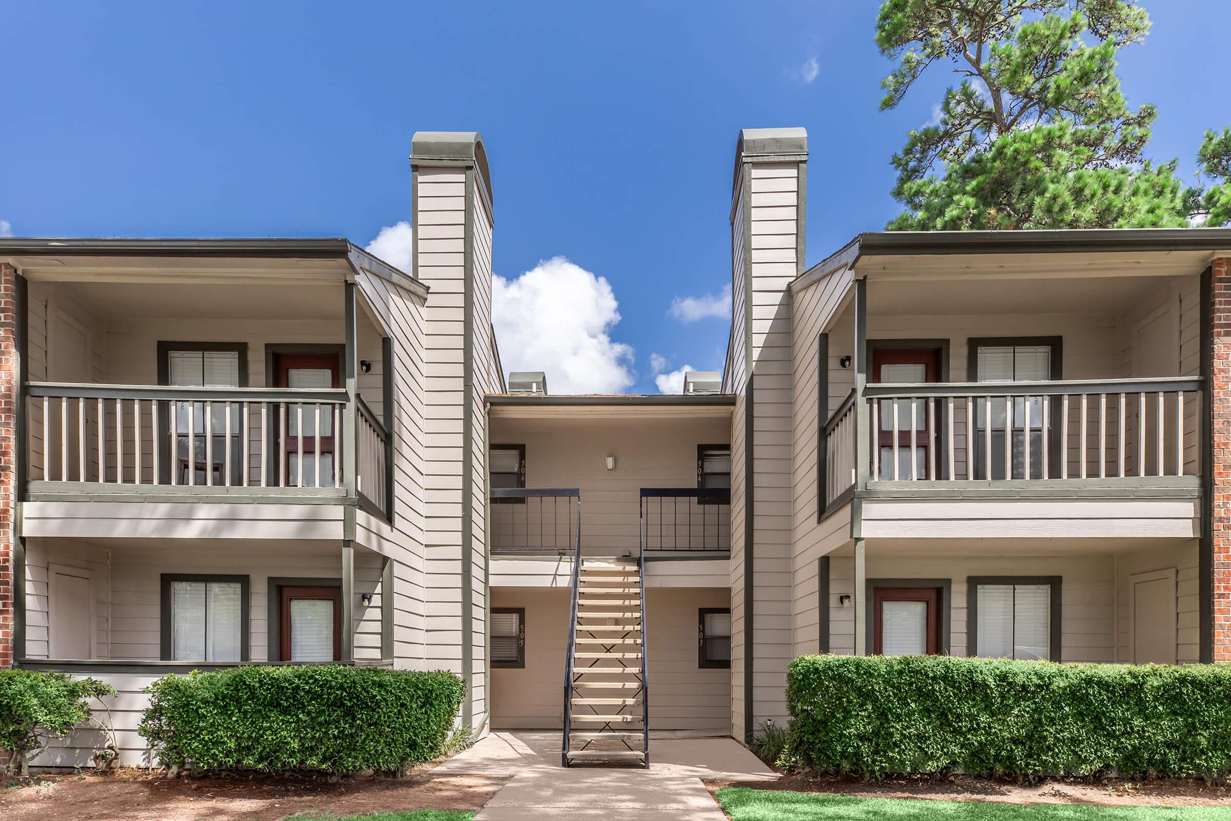 Two-story apartment building with a central staircase leading to the upper balcony. Each unit has a front door and a small balcony, surrounded by well-maintained shrubs and greenery. The sky is clear with a few clouds, creating a bright and inviting atmosphere.