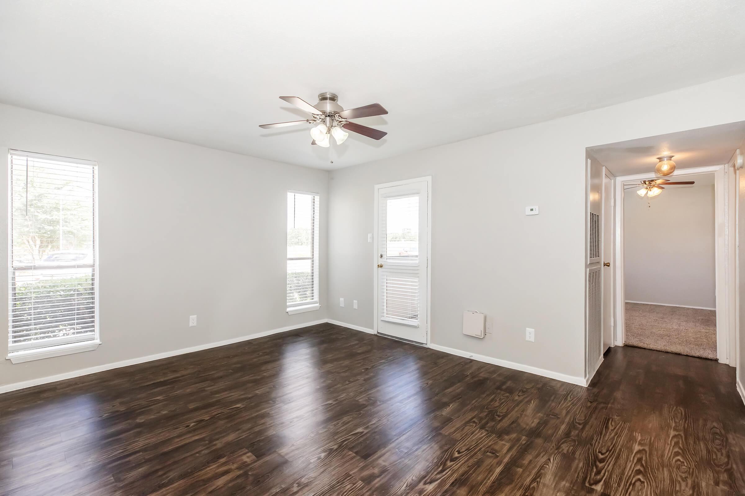 A spacious and well-lit living room featuring hardwood floors, a ceiling fan, and multiple windows. The walls are painted in a light color, and there is a door leading to the outside and a hallway to another room. The overall atmosphere is bright and inviting.
