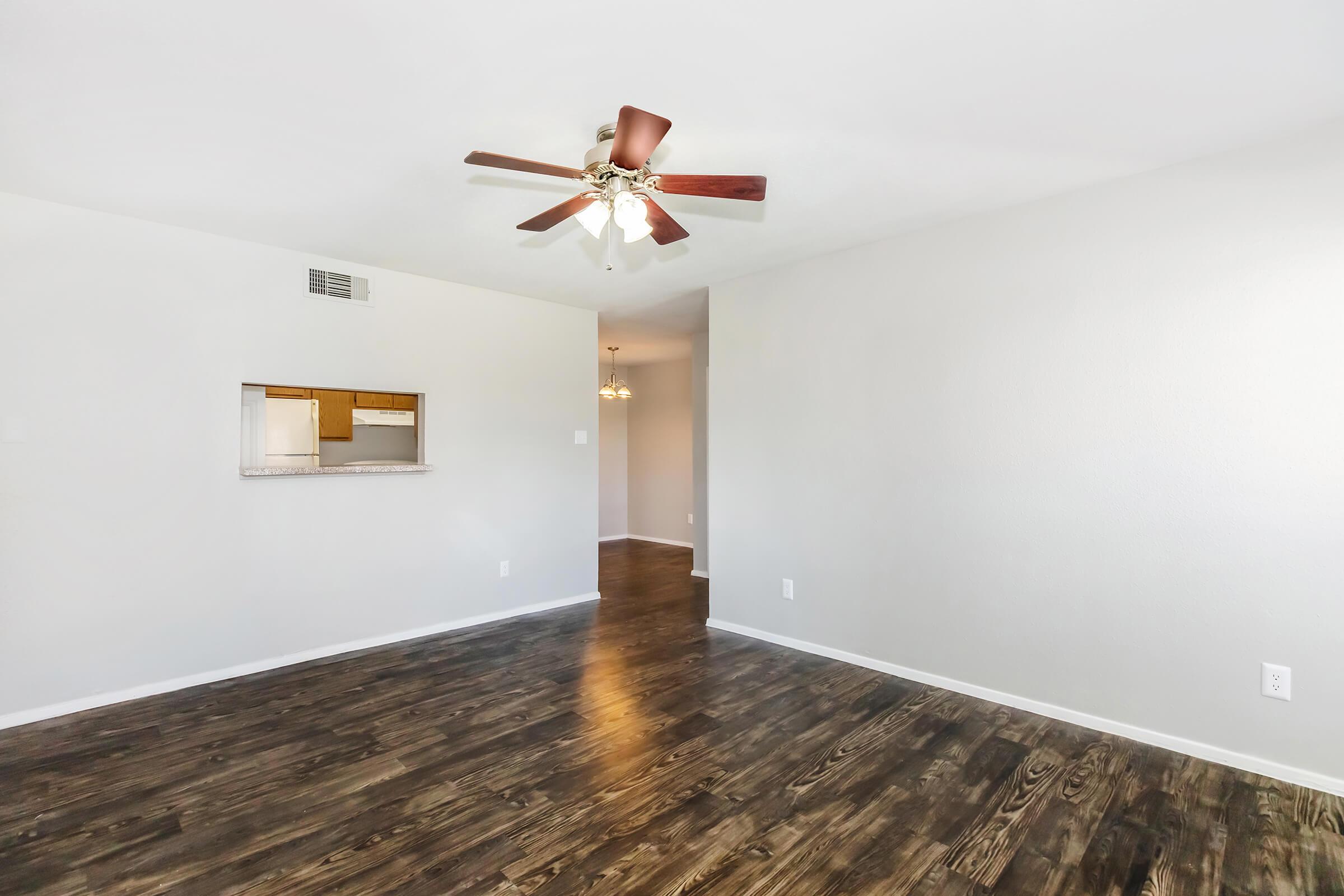 Spacious living area with hardwood-style flooring, a ceiling fan, and a light-colored wall. An open doorway leads to another room, and a small pass-through window opens into a kitchen area. Natural light brightens the space, creating a welcoming atmosphere.