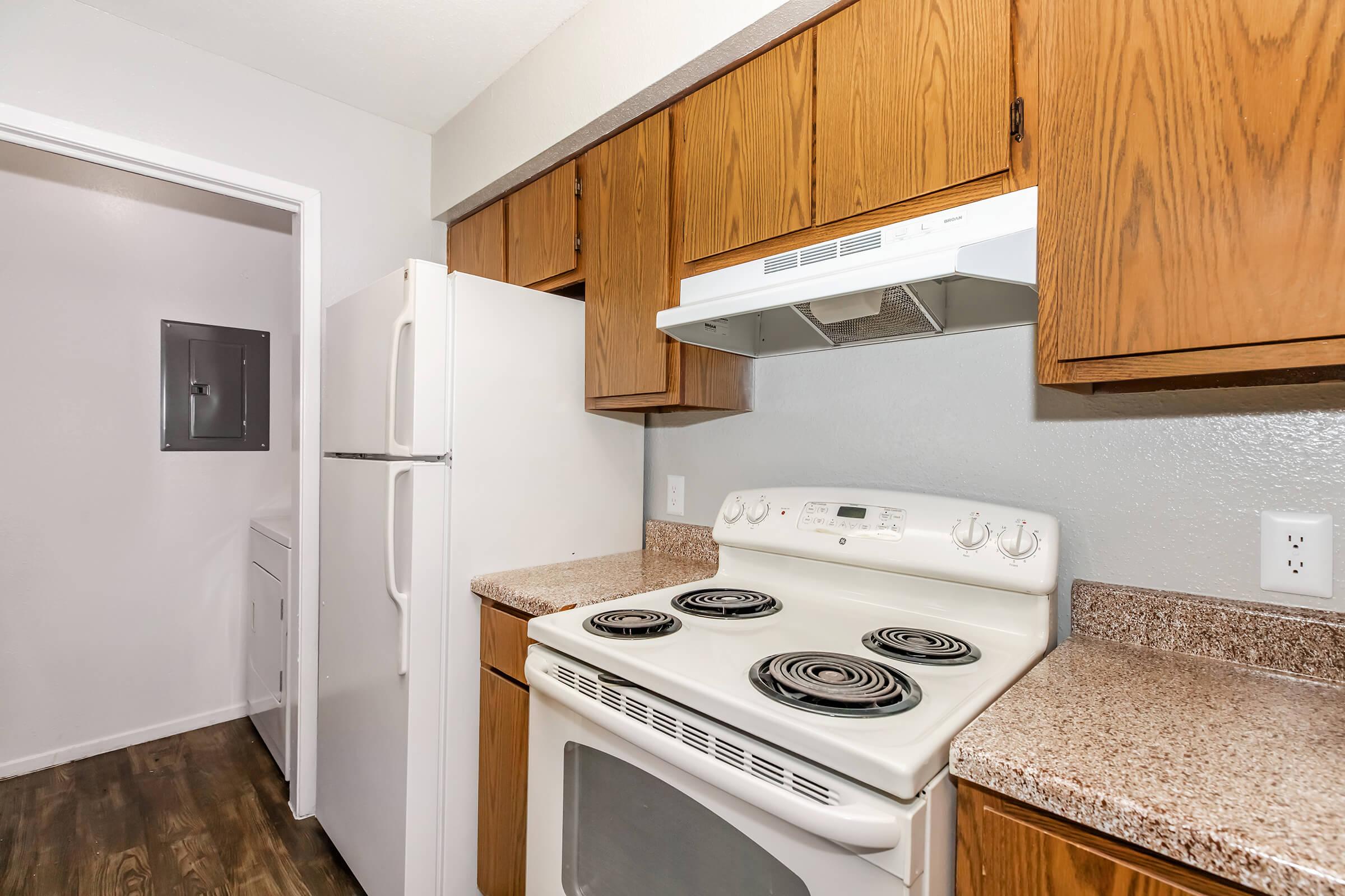 A kitchen featuring a white stove with four burners, a white refrigerator, and wooden cabinets above a speckled countertop. The walls are painted light gray, and there's a doorway leading to a laundry area in the background, with dark vinyl flooring visible.