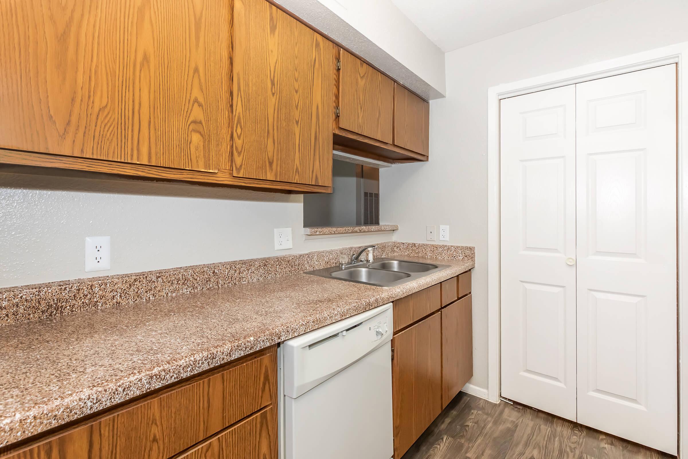 A modern kitchen featuring wooden cabinets, a granite countertop, a double sink, and a dishwasher. There is a mirror above the sink and a closed closet door in the background. The flooring is a dark wood laminate, creating a warm and inviting atmosphere.