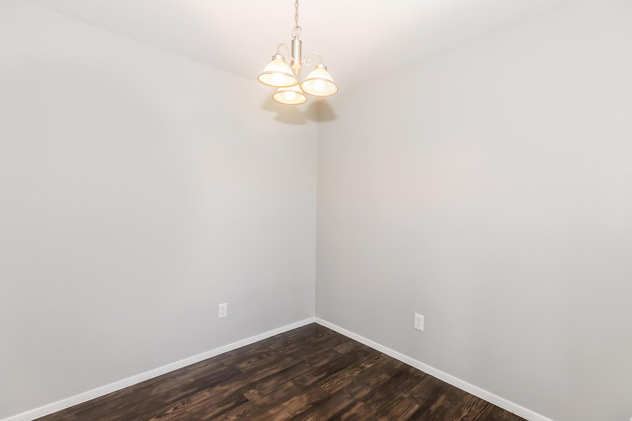 A well-lit empty corner of a room featuring a simple chandelier hanging from the ceiling, light gray walls, and dark wood flooring. The space appears clean and uncluttered, suitable for various uses or decoration.