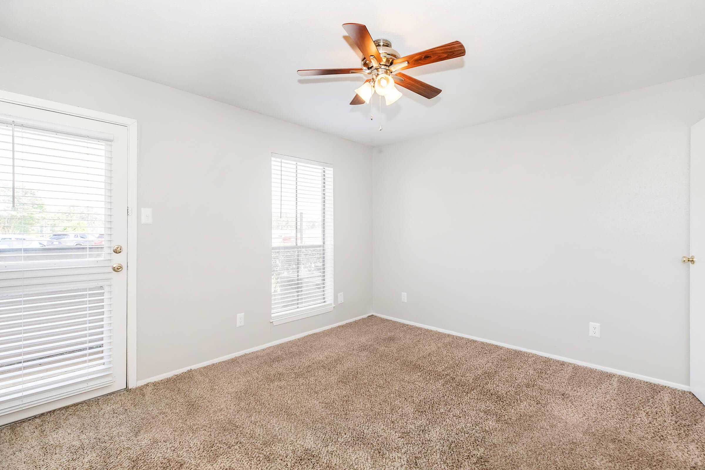 A well-lit room featuring light gray walls and plush beige carpeting. There is a ceiling fan with wooden blades, and two windows with white blinds. A door leads outside, and the overall space appears clean and inviting, suitable for various uses.
