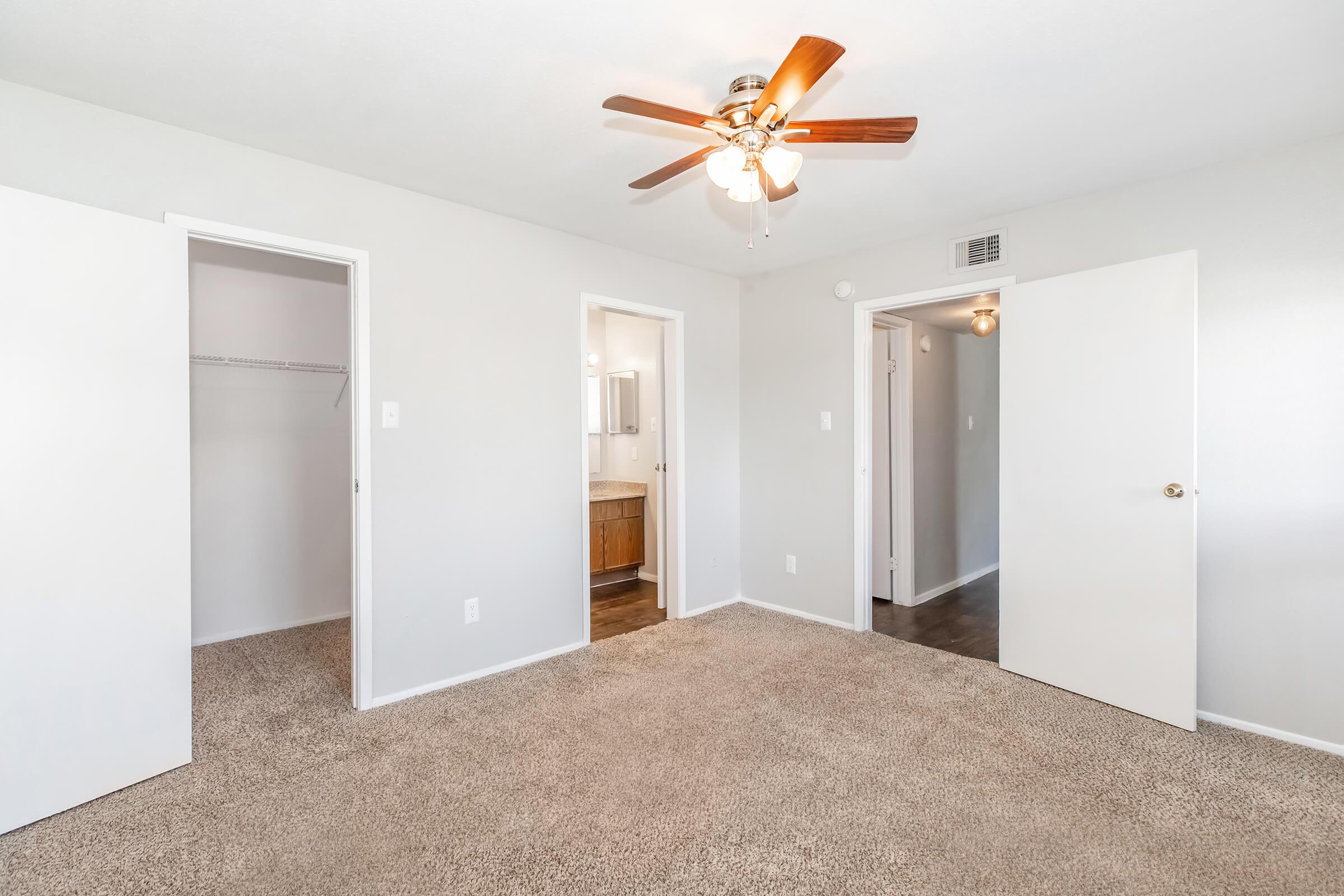 A well-lit bedroom with light gray walls and plush carpet. It features a ceiling fan, two open doors leading to a closet and a bathroom, and a spacious layout that suggests an inviting atmosphere. The room is empty, making it adaptable for various furniture arrangements.