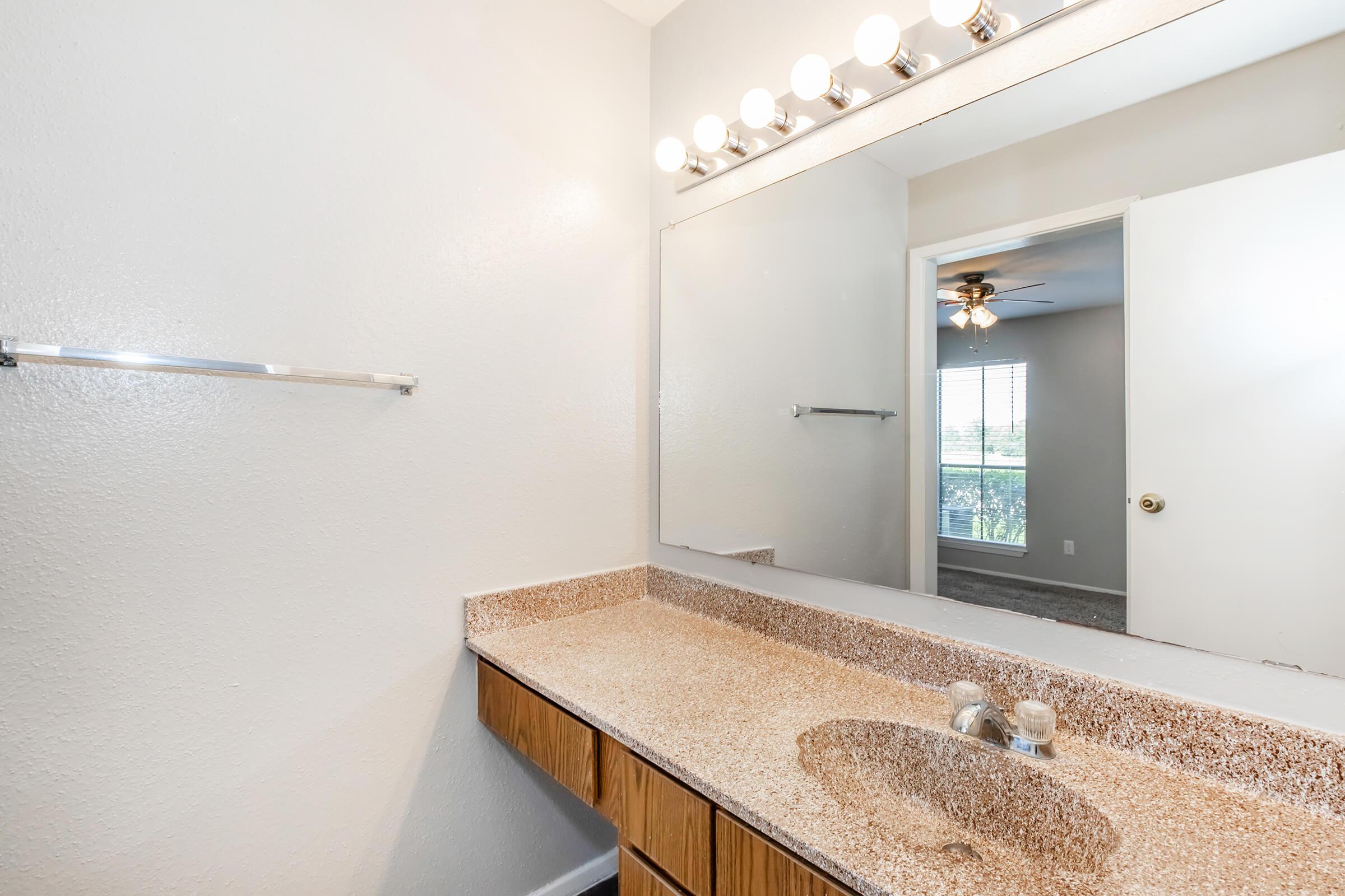 A bathroom vanity with a lighted mirror above a speckled countertop, featuring two faucets. An empty towel rack is mounted on the wall. In the background, a doorway leads to another room, partially visible with a ceiling fan and window. The walls are painted in a light color.