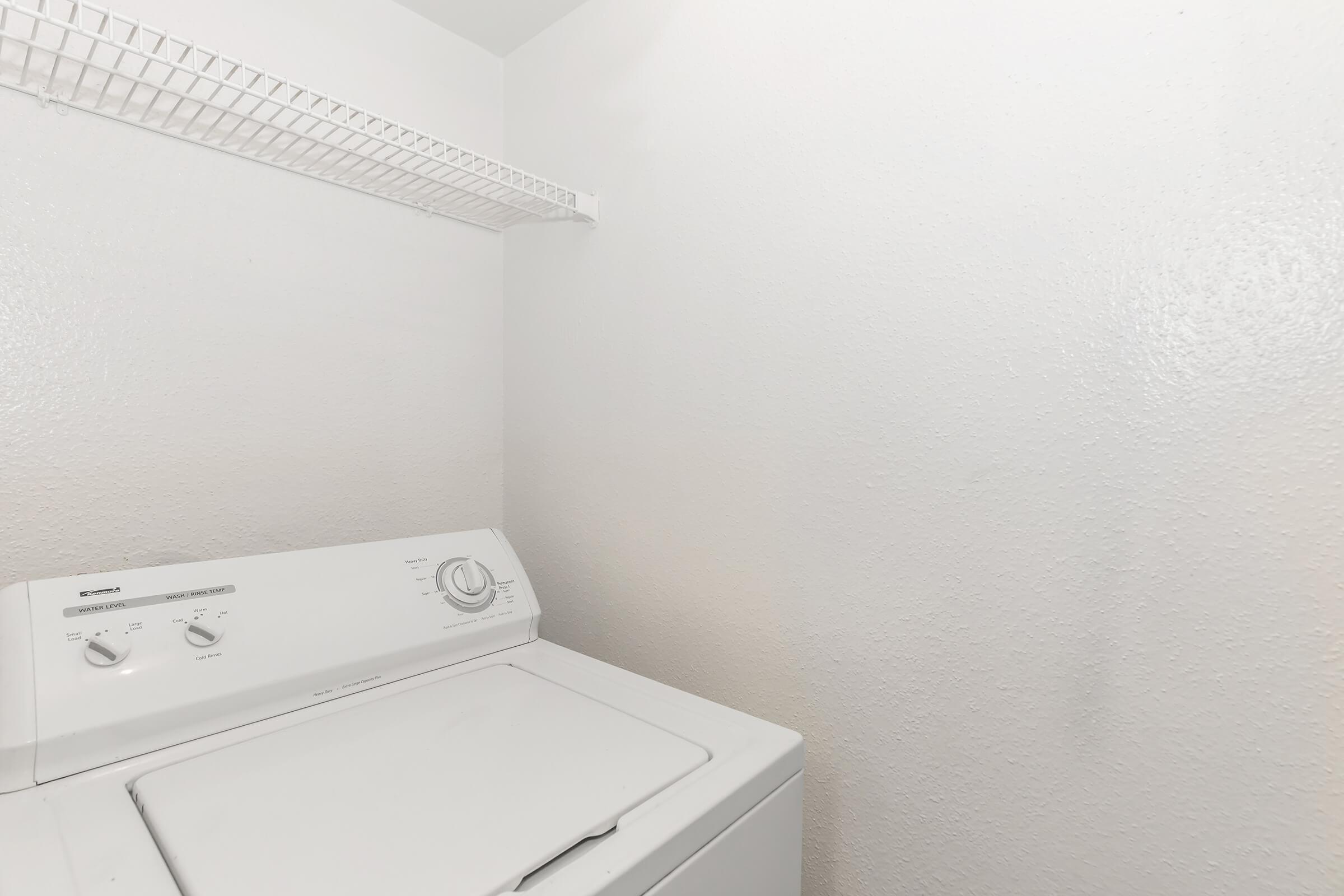 A clean, bright laundry room featuring a white washing machine against a plain white wall. A wire shelf above provides storage space, enhancing the functional and minimalist design of the area. The room appears well-maintained and organized.