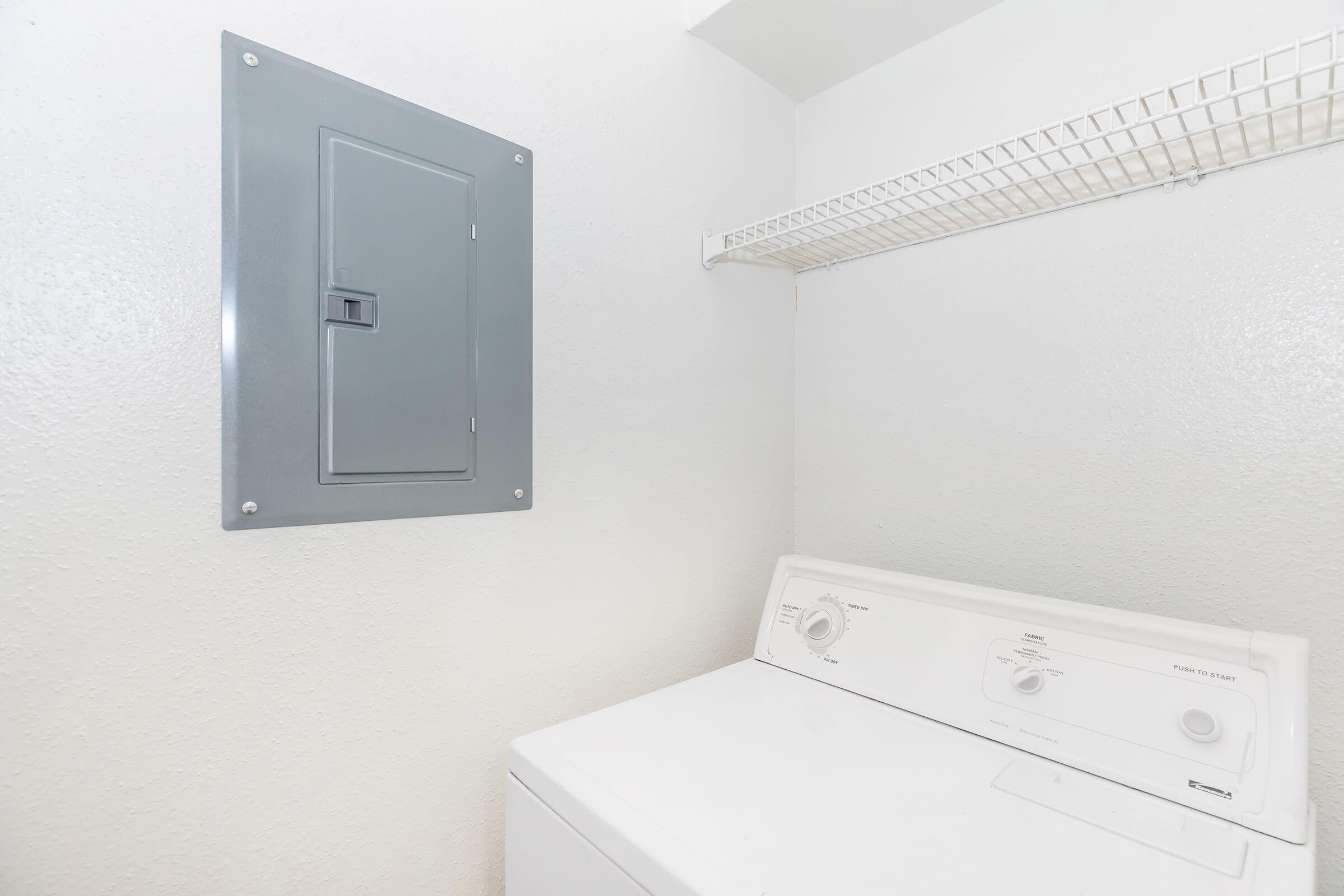 A clean laundry room featuring a white washing machine and a gray electrical panel mounted on a white wall. A wire shelf is visible above the washing machine, providing additional storage space. The overall ambiance is bright and uncluttered.