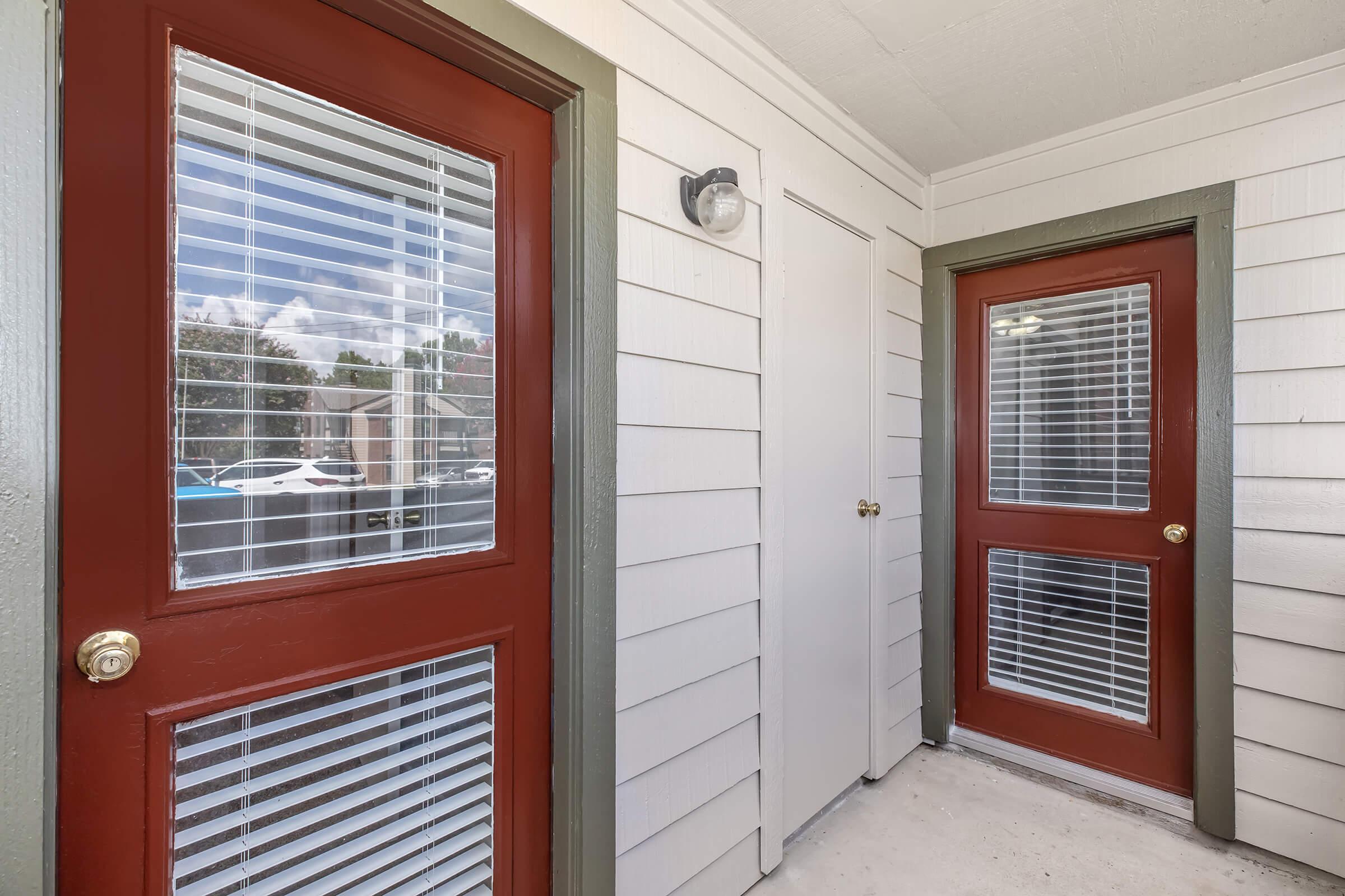 Exterior view of a porch featuring two doors with red frames and white blinds. The walls are light-colored wood siding, and there's a light fixture above one of the doors. A glimpse of outdoor scenery is visible through the windows, suggesting a residential setting.