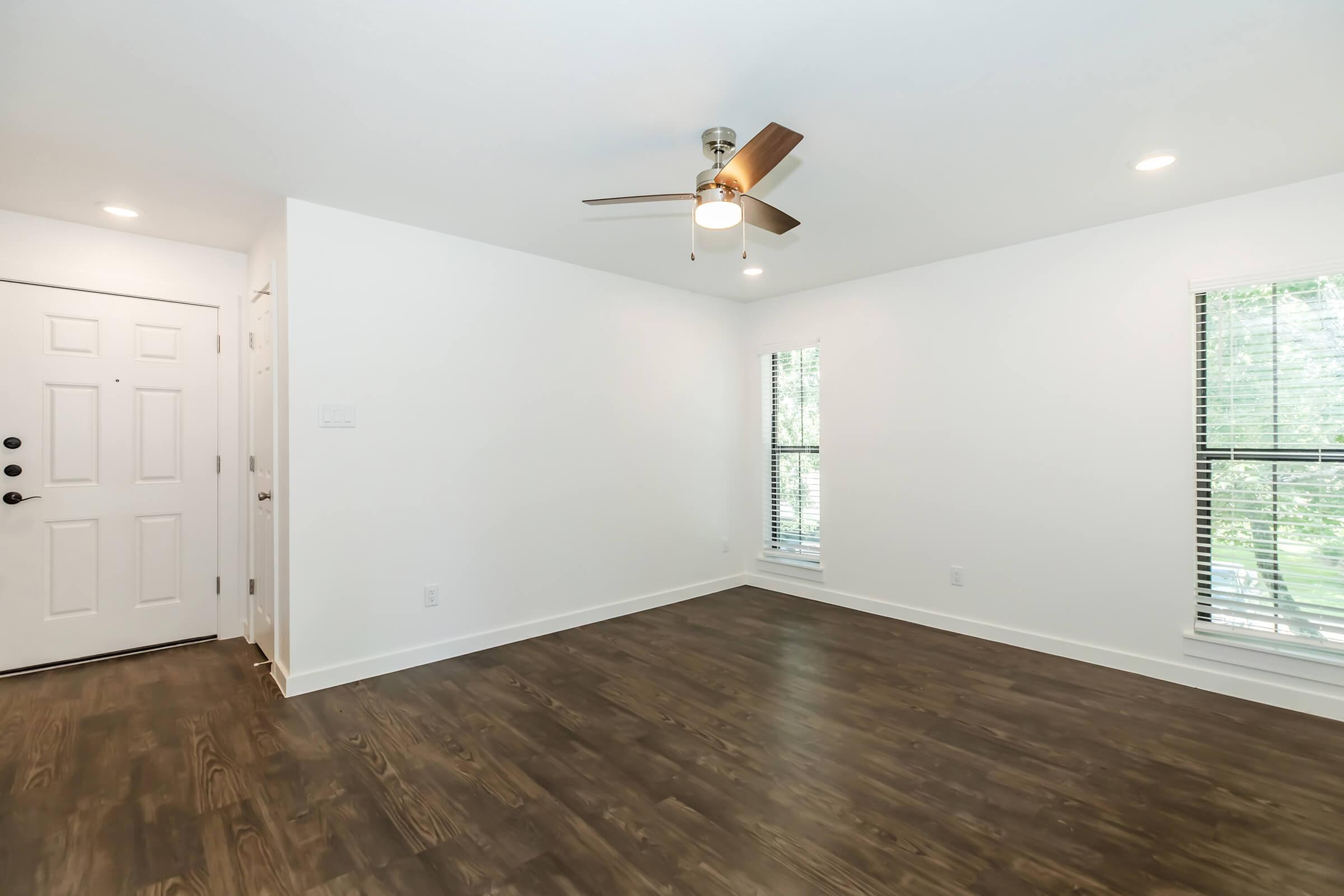 Bright interior of a room featuring light wooden flooring, a ceiling fan, and two windows with blinds. The walls are painted white, and a door is visible on the left side. The space is empty and well-lit, providing a sense of openness and potential for decoration.