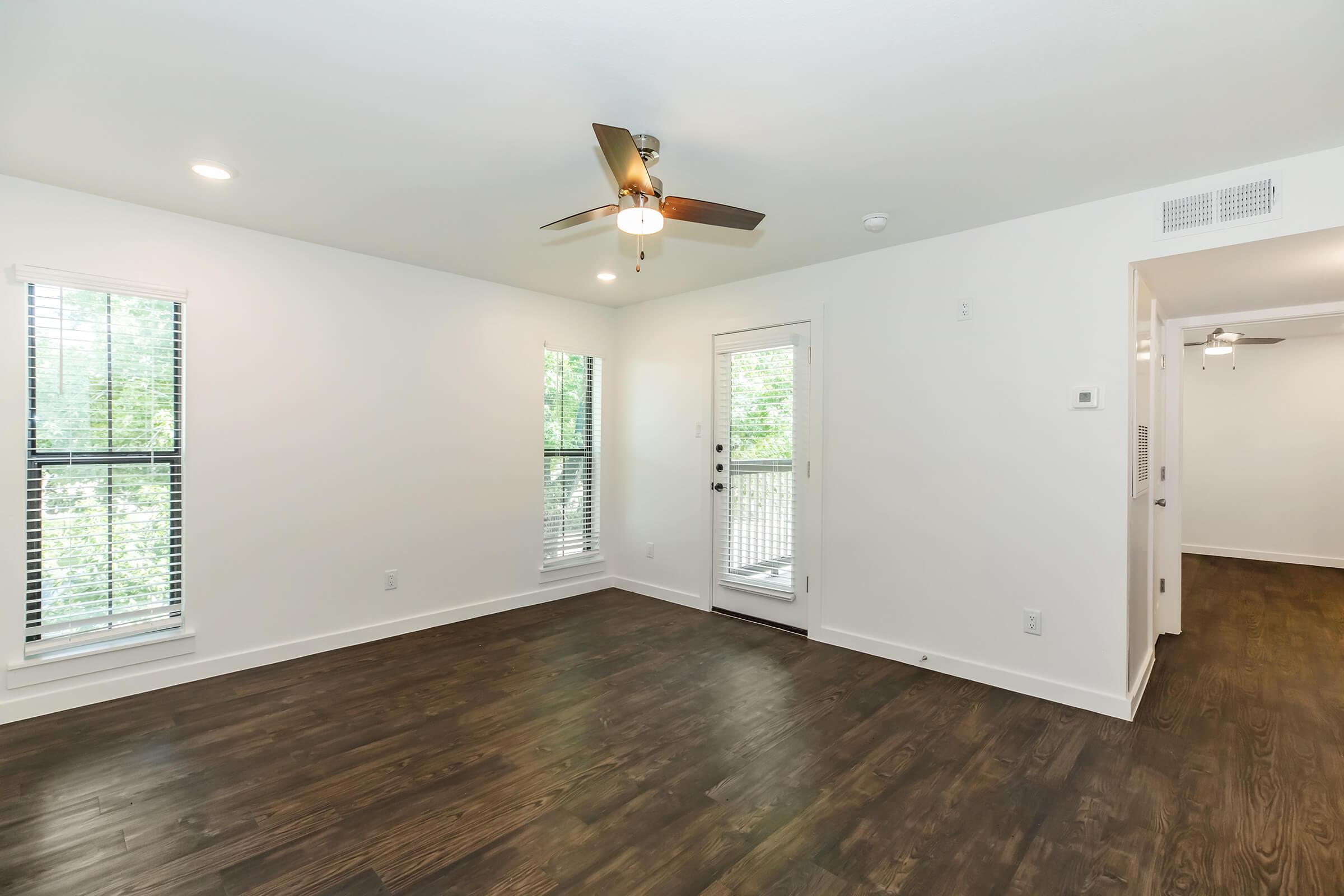 A bright, open living space with a ceiling fan and hardwood flooring. There are two windows with blinds allowing natural light, and a door leading to a balcony or patio. The walls are painted white, creating a clean and modern atmosphere, while adjacent space is visible through an open doorway.