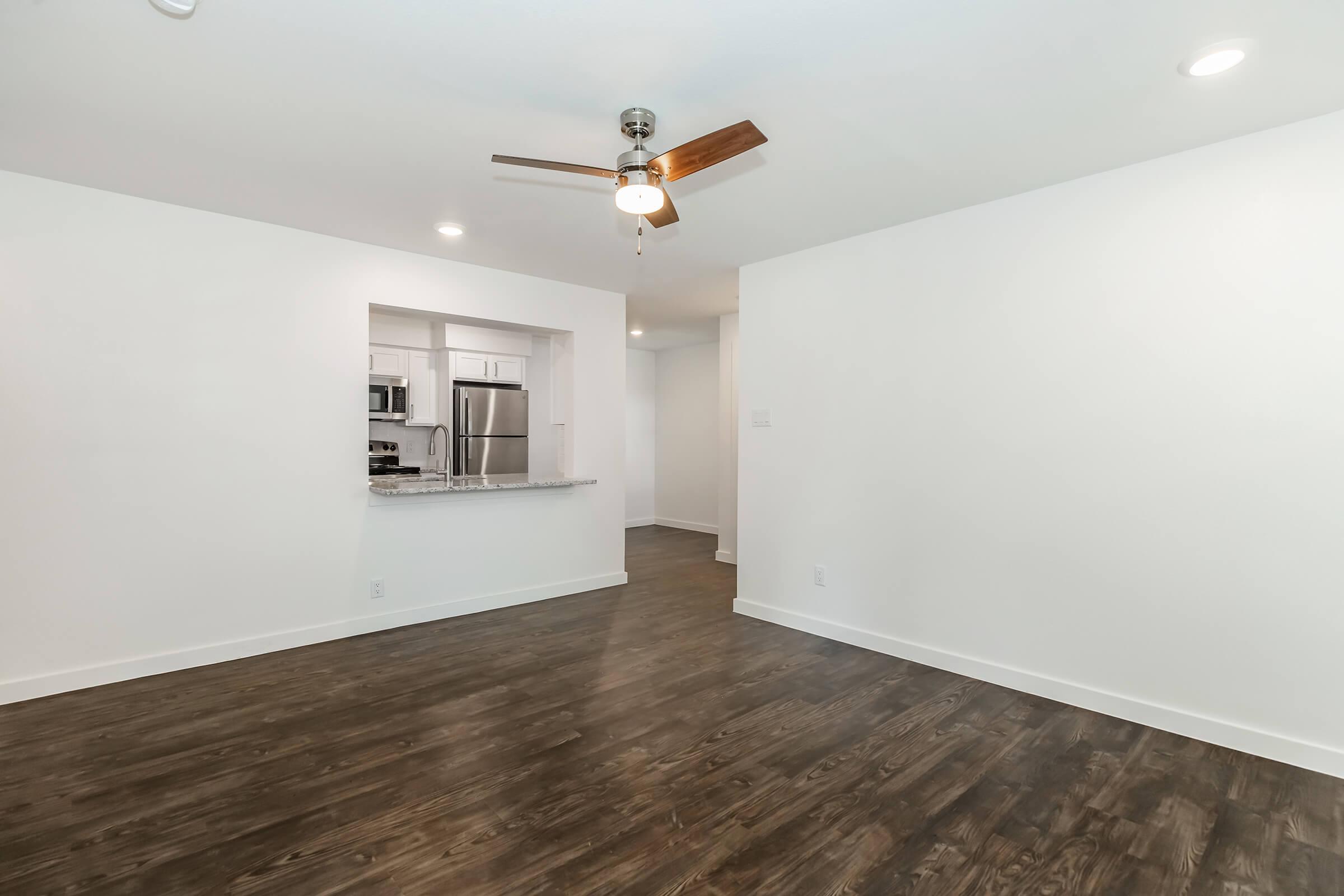 A spacious and bright living area with wood-like flooring, a ceiling fan, and a modern design. The room features white walls and a view of a kitchen area with stainless steel appliances and a granite countertop, creating an open and inviting atmosphere.