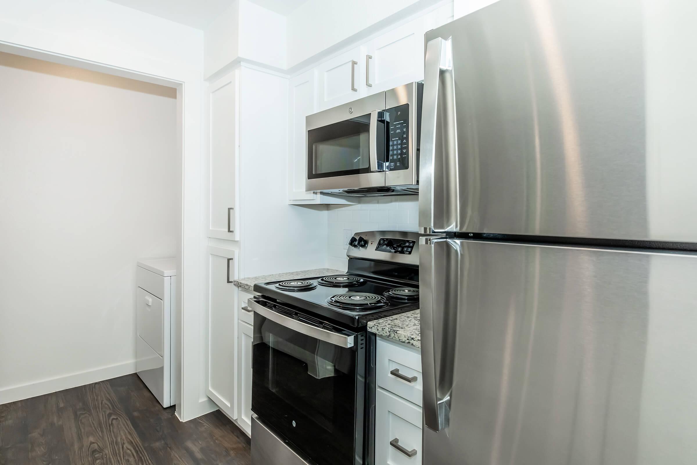 A modern kitchen showcasing stainless steel appliances including a refrigerator, microwave, and stove. The cabinets are white with a granite countertop. A washer and dryer are partially visible in an adjacent space, and the floor is dark wood. Overall, the design is clean and contemporary.