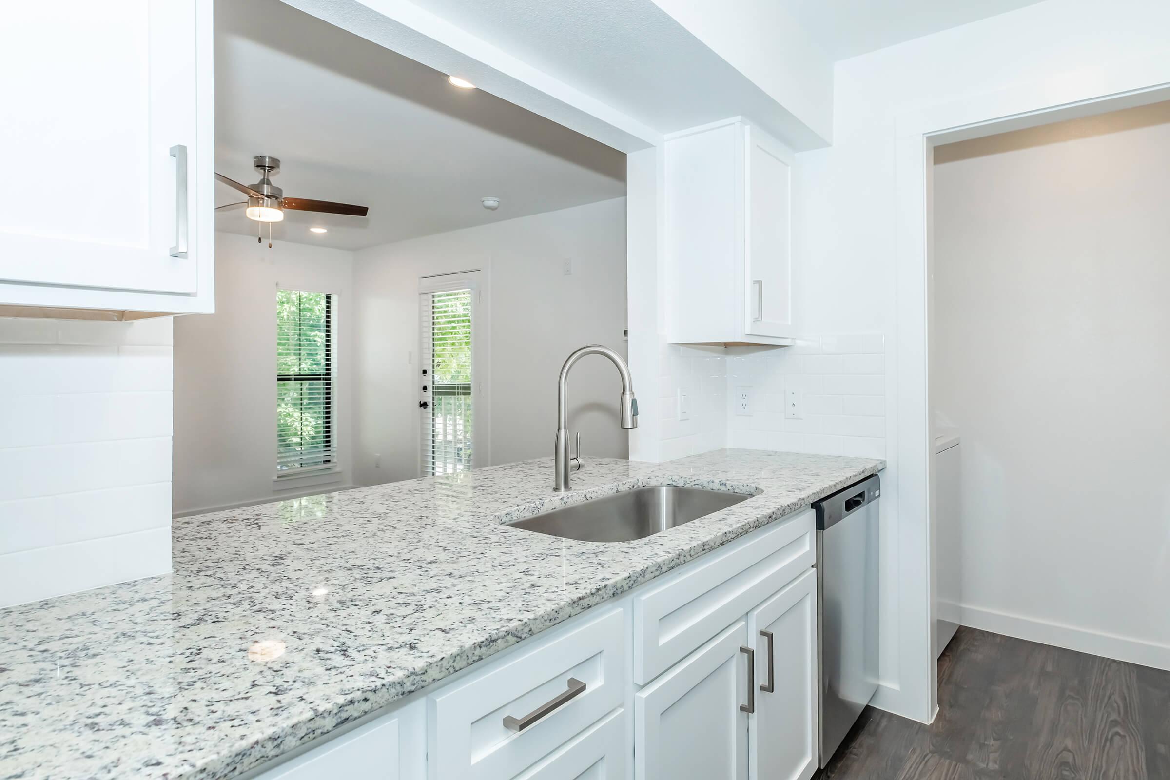 A modern kitchen with a granite countertop, stainless steel sink, and white cabinetry. A ceiling fan is visible, with natural light coming through a window. The background shows a living area and a doorway leading to another room. The overall design is bright and contemporary.