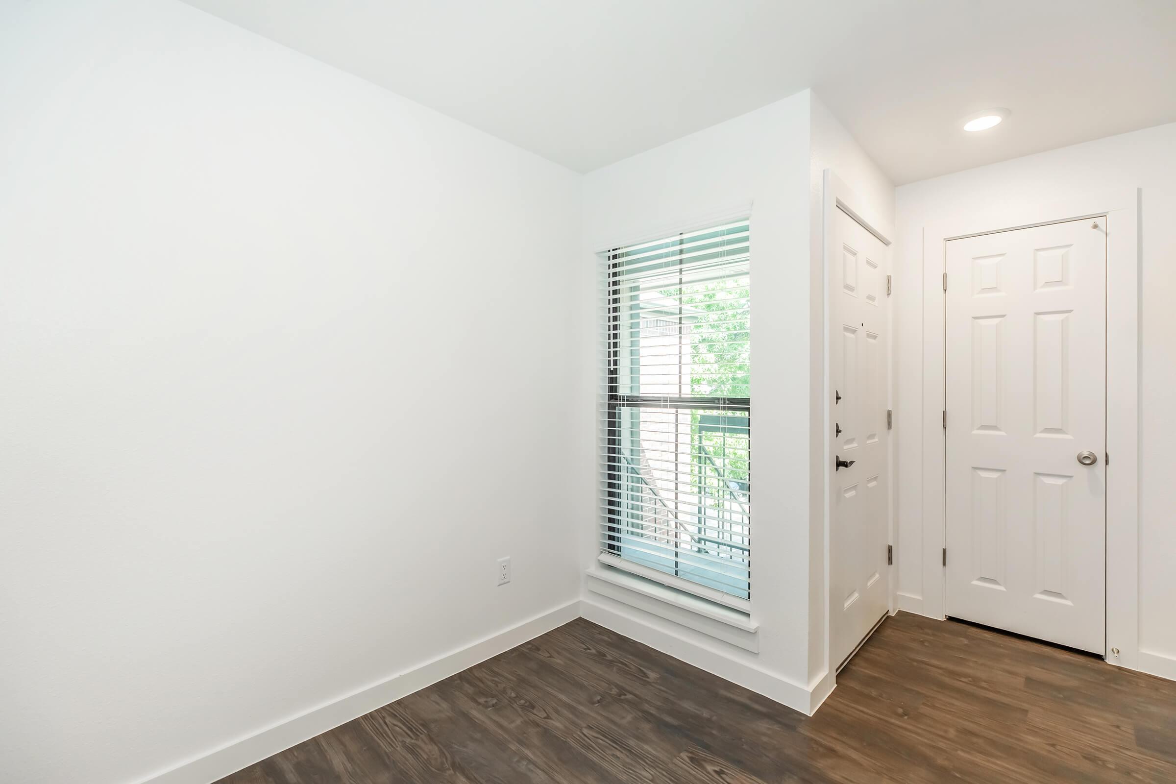 A bright and spacious room featuring white walls, wooden flooring, and a window with blinds. There is a doorway leading to another room and a closet door visible. The area is well-lit and has a clean, modern aesthetic.
