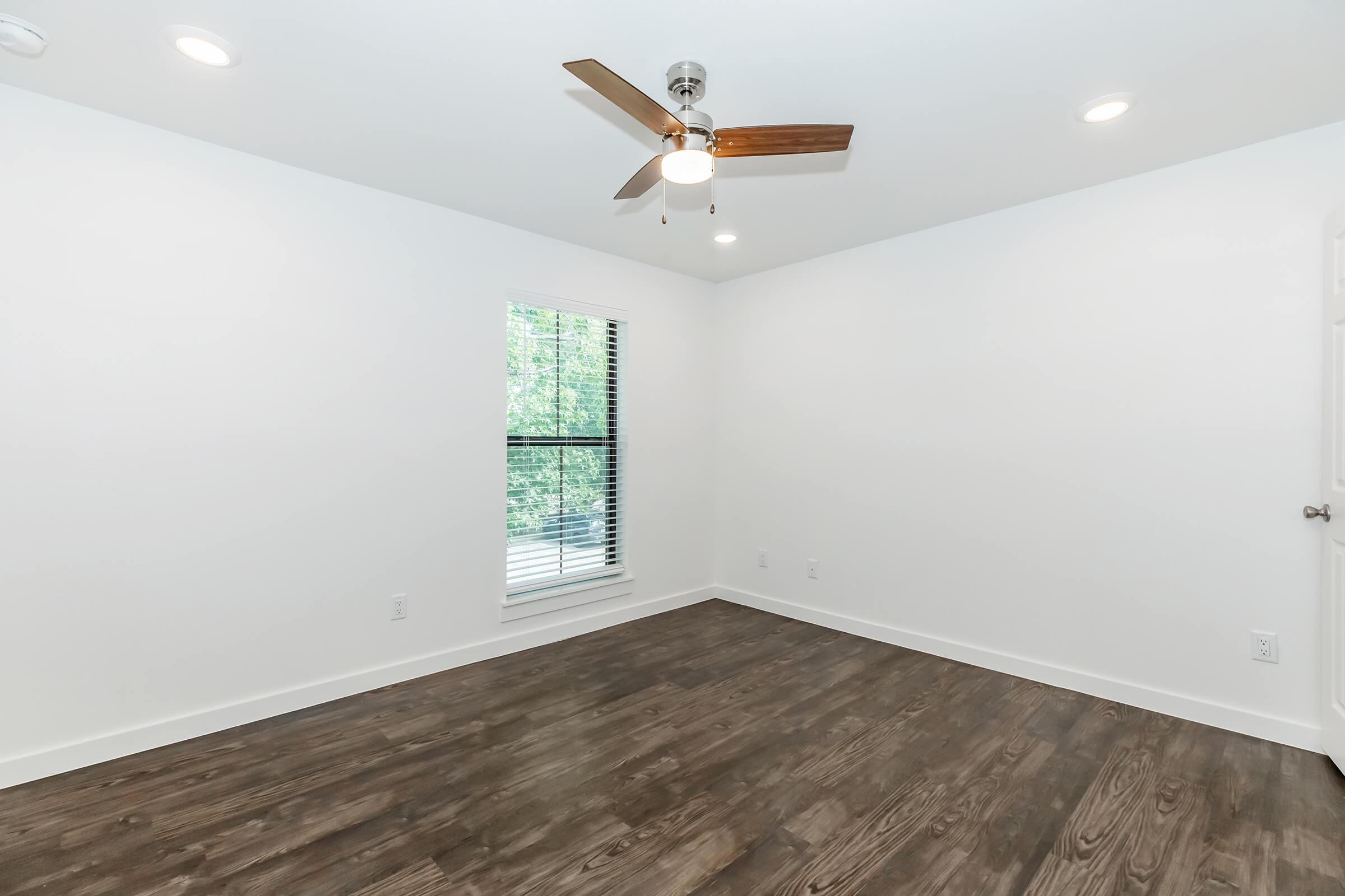 A bright, empty room featuring a ceiling fan, white walls, and a large window with blinds. The floor is made of dark wood, creating a modern and clean aesthetic. Natural light filters in from the window, enhancing the spacious feel of the room.