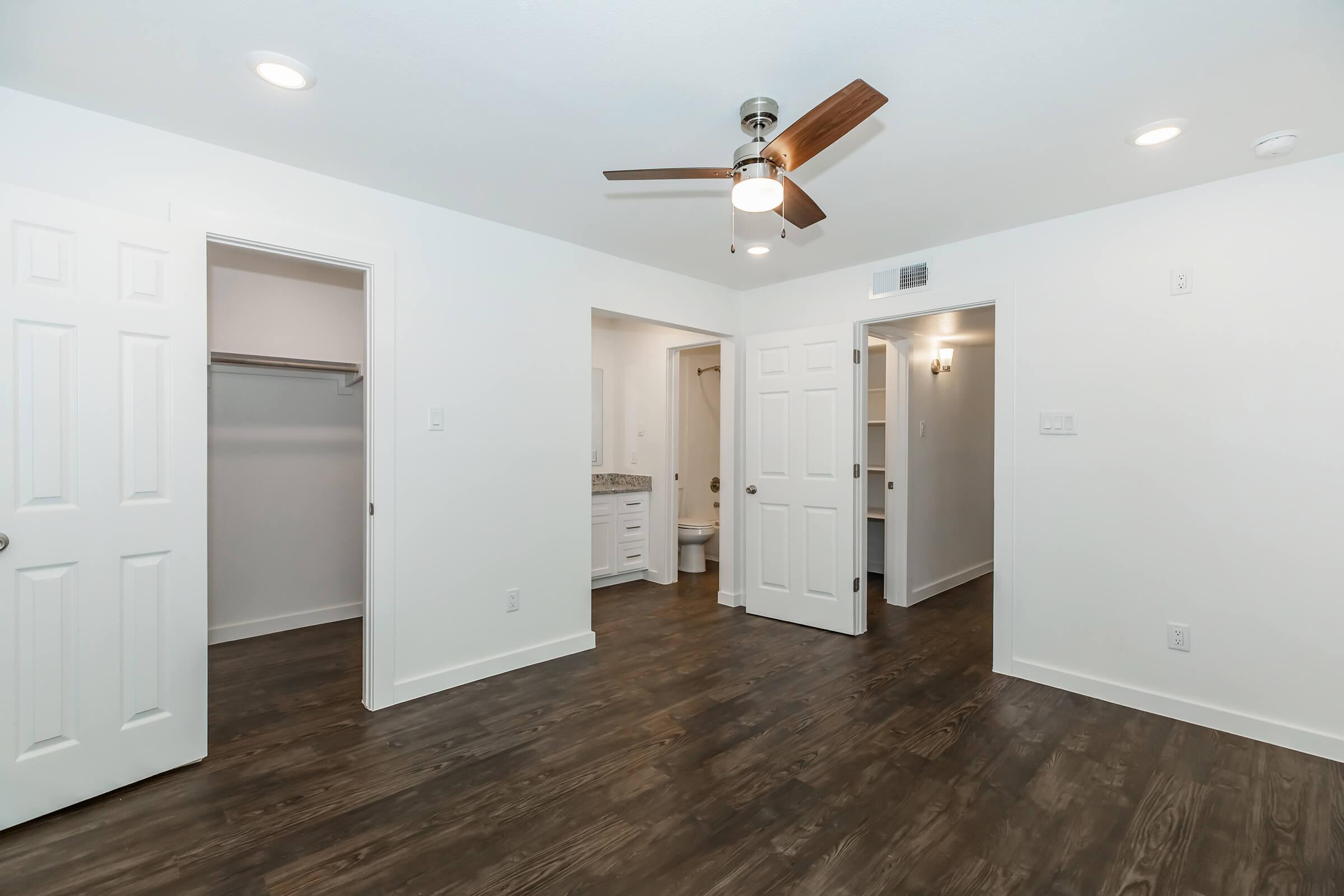 A spacious, well-lit room with dark wood flooring, featuring a ceiling fan. There are two white doors leading to a closet and a bathroom. The walls are painted white, enhancing the brightness. A small vanity area is visible in the distance, adding to the room's functionality.